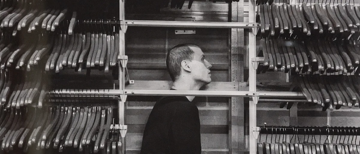 A black-and-white photo of a man with a shaved head standing in profile between two rows of empty hangers on racks. The setting appears industrial and somber.
