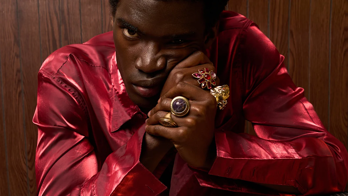 A man looks at the camera. He wears a silk pink shirt and raises his hands up into clenched fists. 