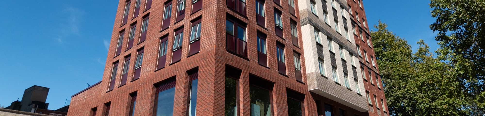 The exterior of a tall modern student accommodation building with a red brick and cream facade, featuring large windows with dark burgundy frames, shot from street level against a clear blue sky with a tree visible to the right.