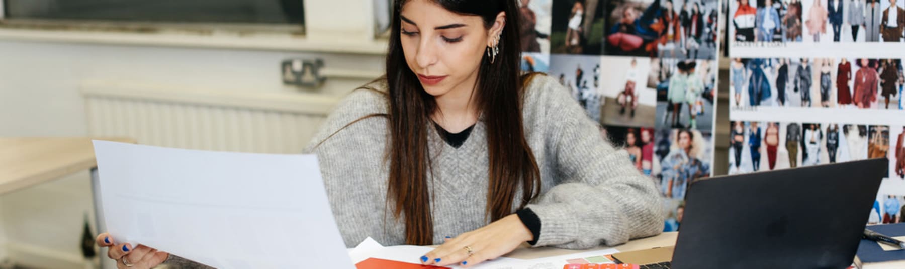 A student sitting at a desk with various pieces of work and a laptop, looking closely at an A3 sheet of paper