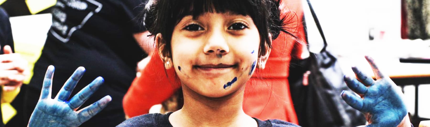 A Young girl holds up her hands showing that they are covered in blue paint, she also has a few paint marks on her face.