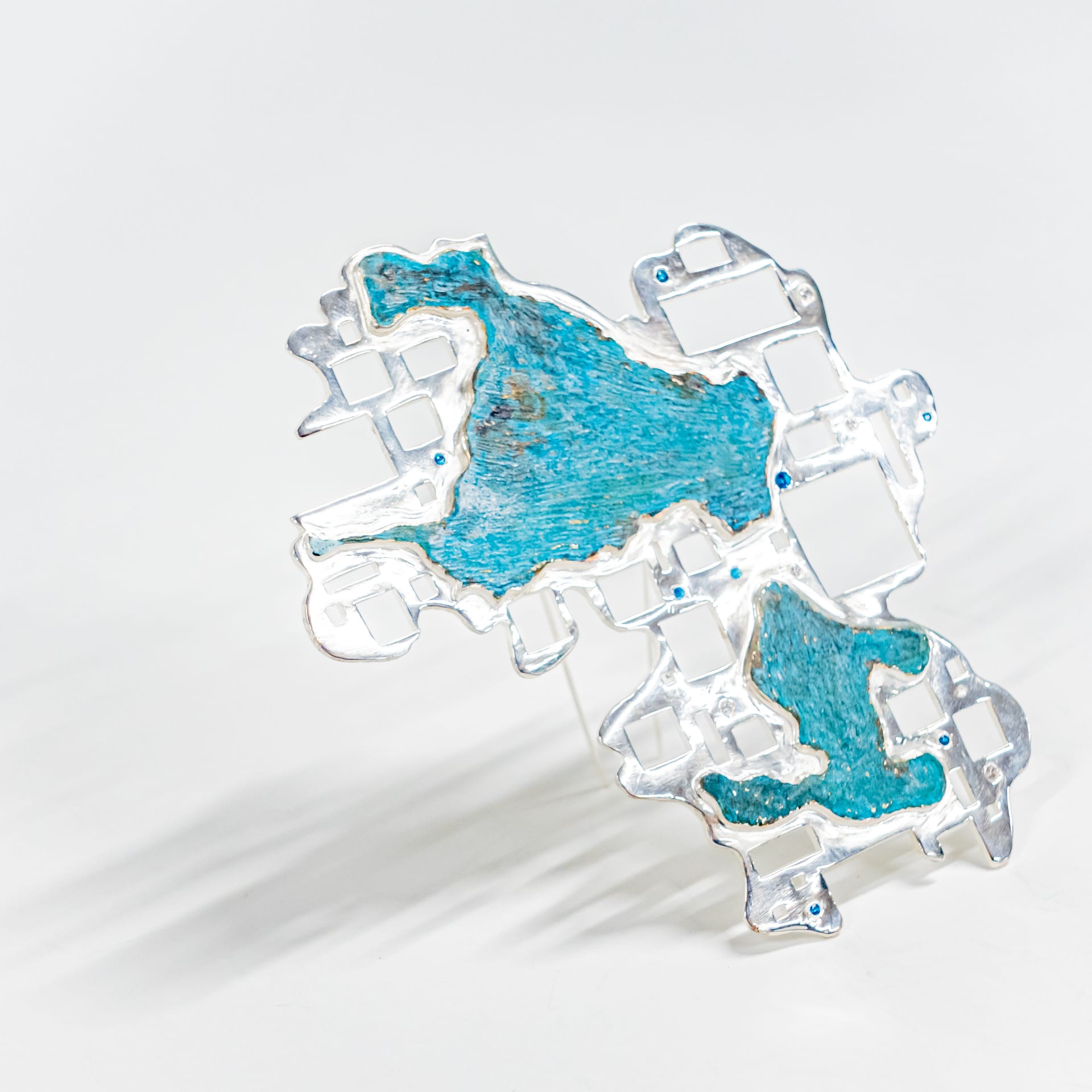 An irregular brooch composed of a silver-coloured lattice frame with copper-green filling (symbiotic), embellished with blue zirconia.