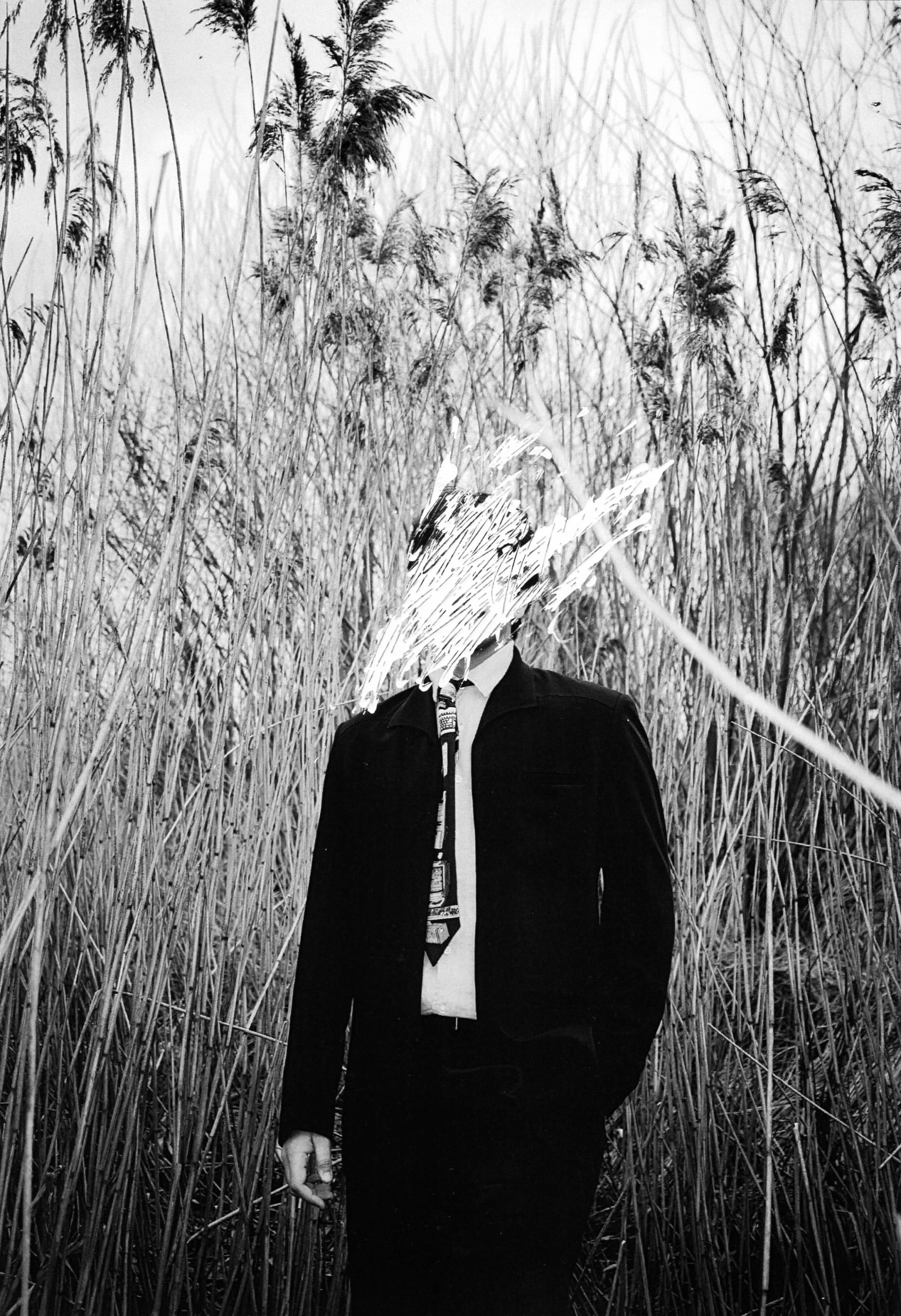 A man stands in a long grass field with his face scribbled out.