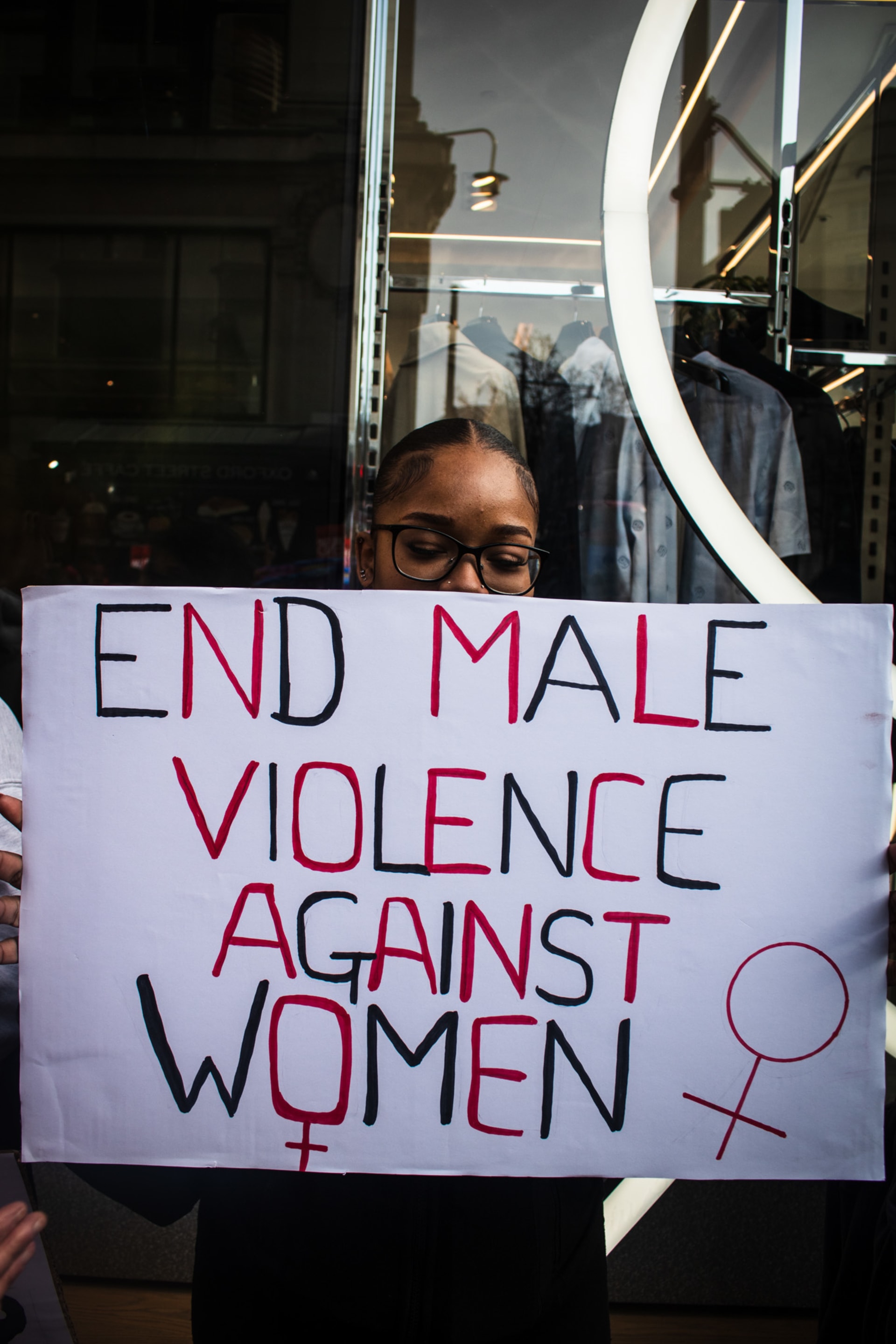 A young woman holding a handmade sign saying: " END MALE VIOLENCE AGAINST WOMEN " in all capitals in black and red lettering. 