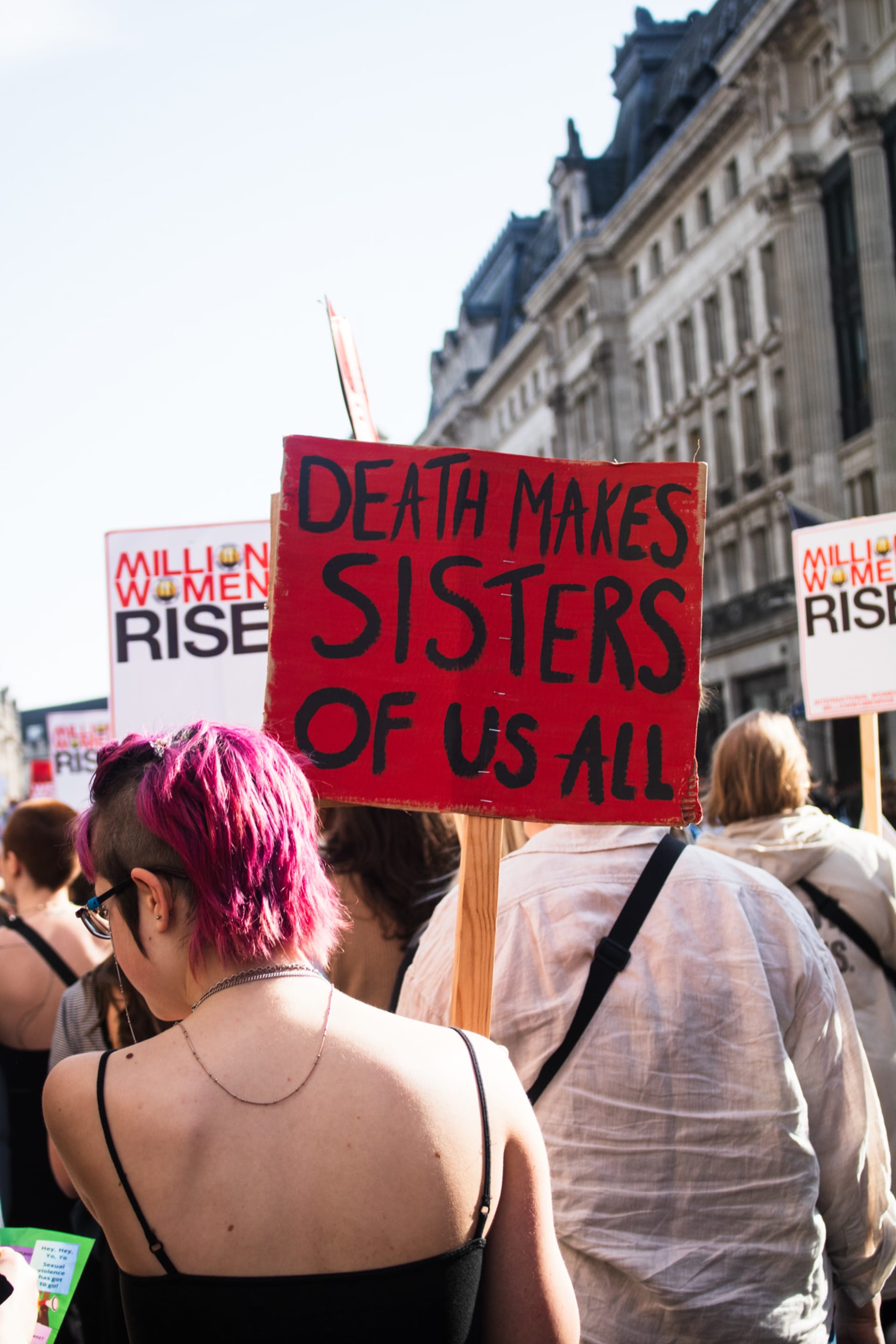 Image of a march participant with pink hair from behind them, holding a hand painted sign saying: "Death makes sisters of us all" In black lettering 