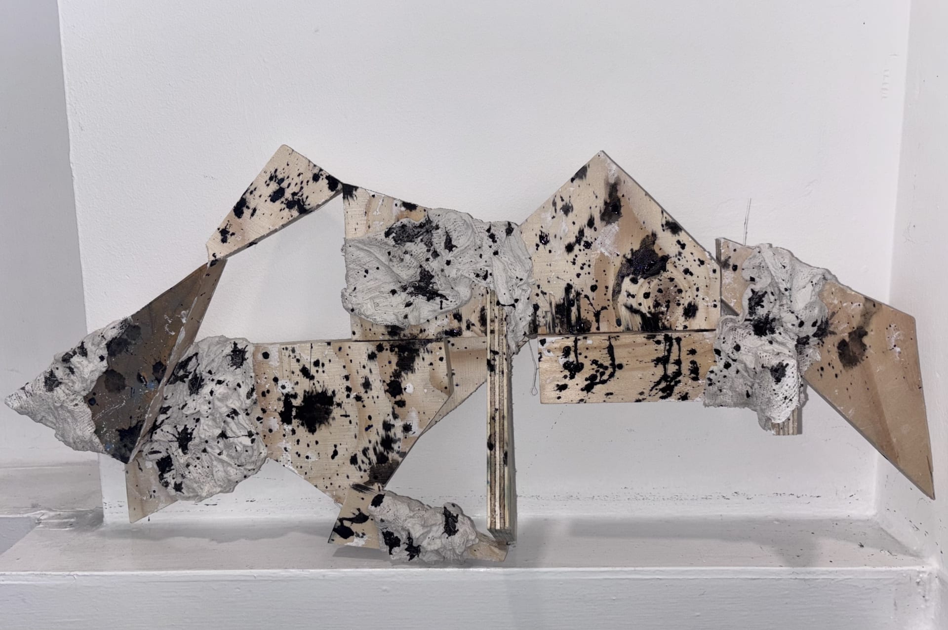 Triangle shaped wood pieces are joined together in an abstract composition and covered with thick plaster, then splattered with black paint.