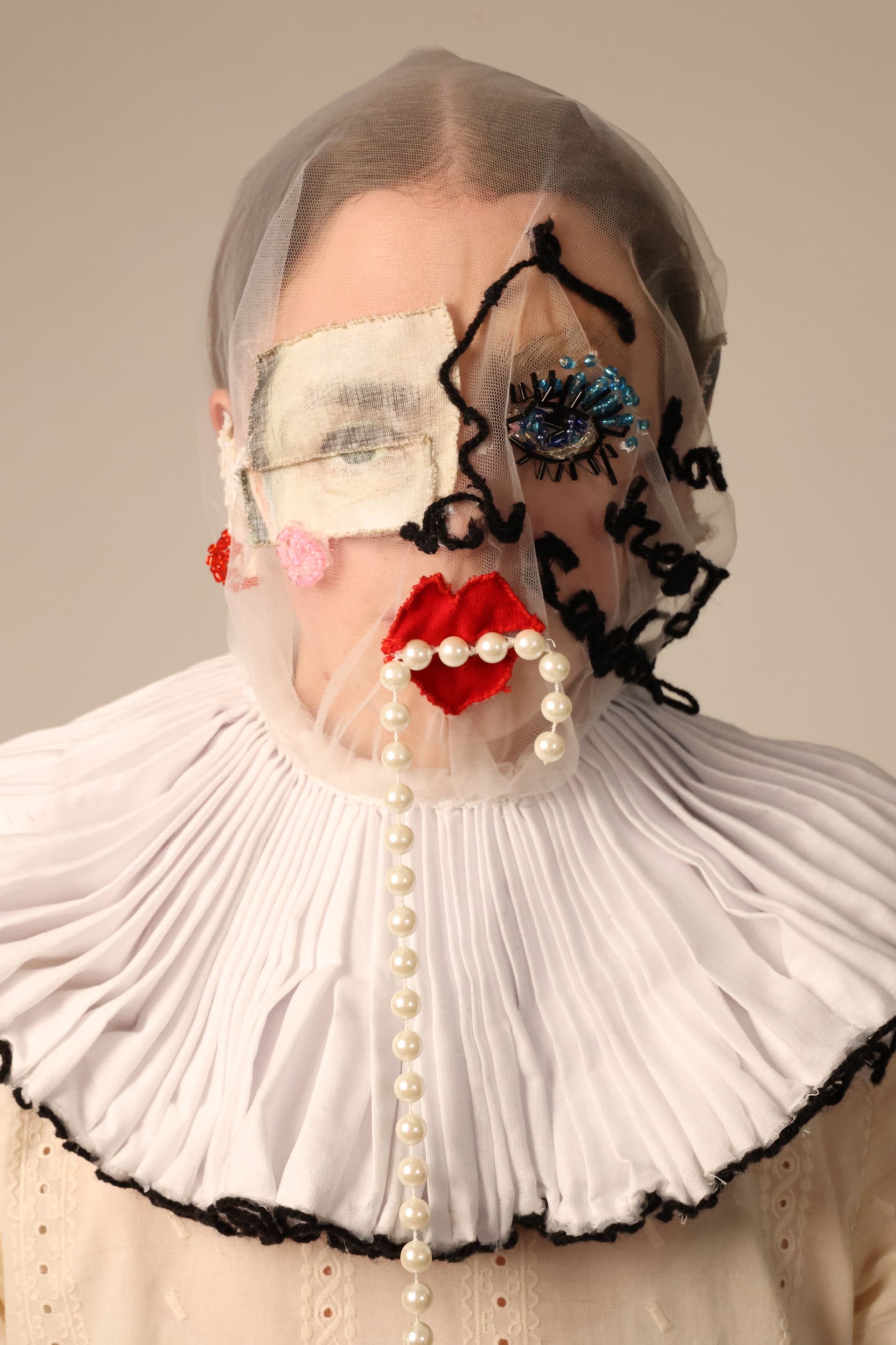 Headshot of person wearing mesh mask with a ruff collar. The mask contains facial features made from fabric, beads, wool, and acetone printed fabric.