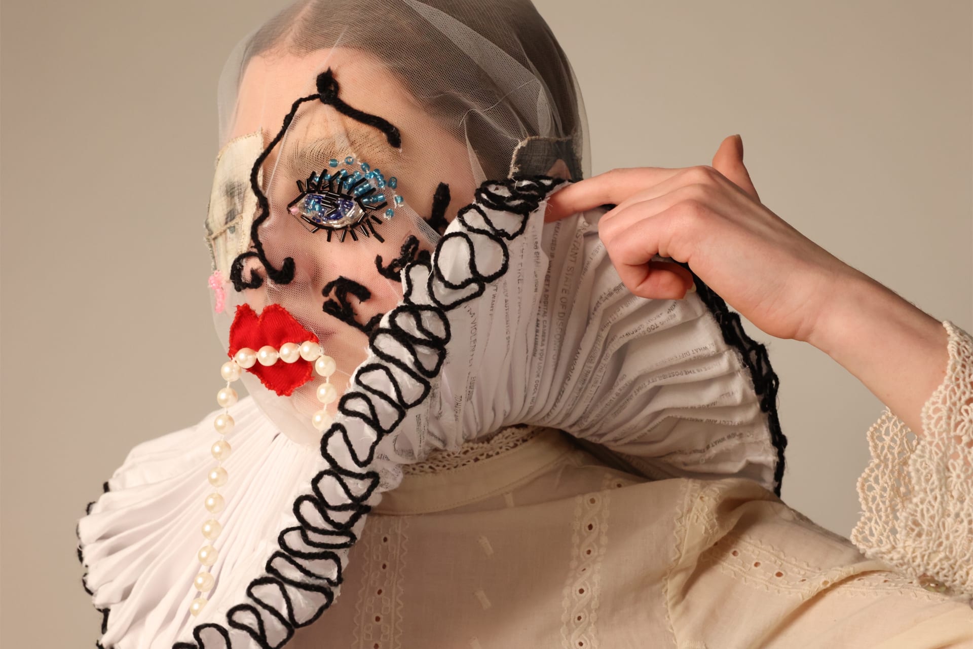 Mesh mask with sewn features with ruff collar. The ruff is being lifted to reveal strips of text underneath - not all text is visible or clear.