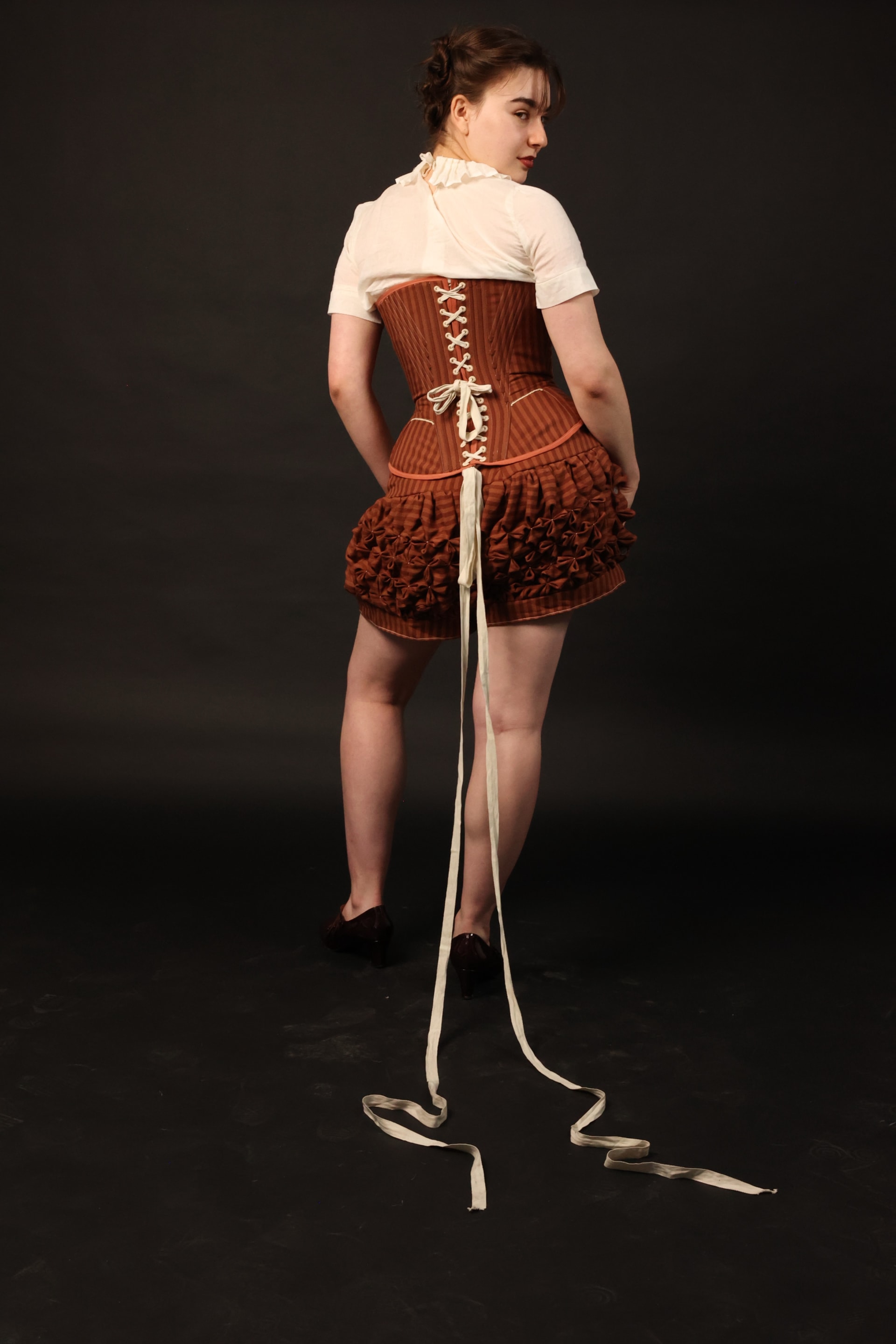 Model with body facing backwards, wearing striped shorts , a white blouse/ chemise and a striped corset with the laces trailing behind them.