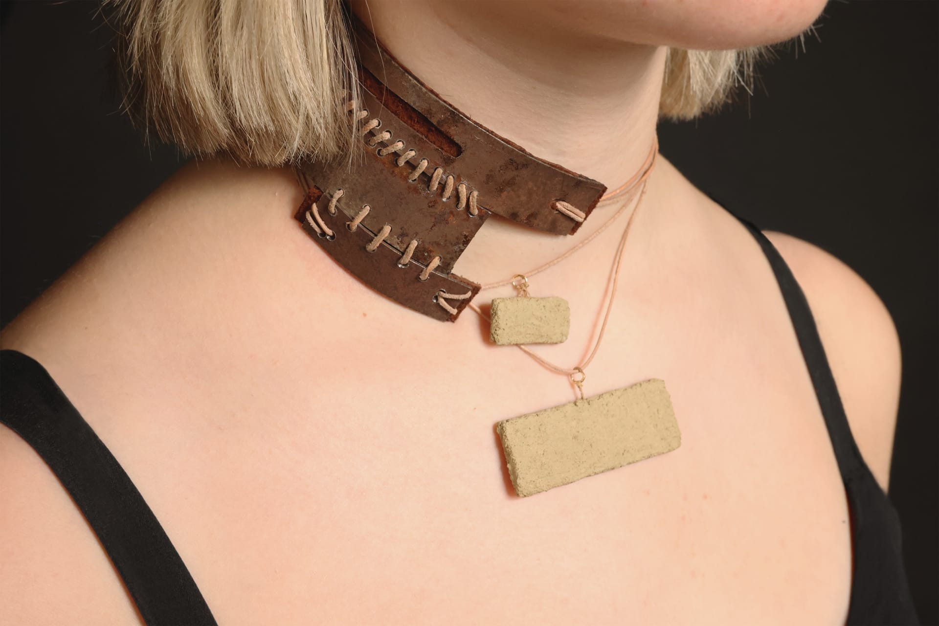 A neck piece made from rusted metal, sewed and held up by a leather cord, from which cement pendants hang.