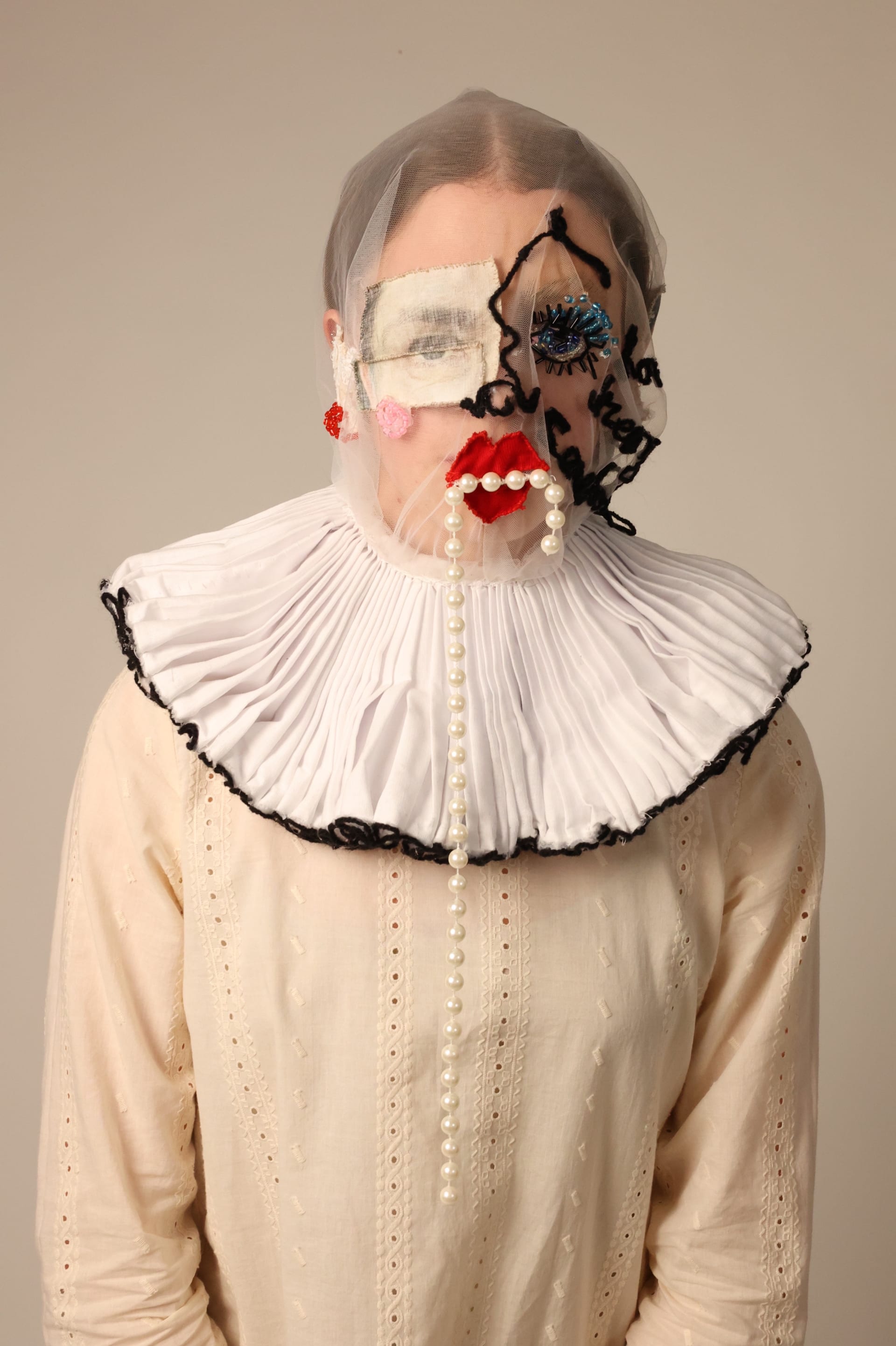Person wearing mesh mask with a ruff collar. The mask contains facial features made from fabric, beads, wool, and acetone printed fabric.