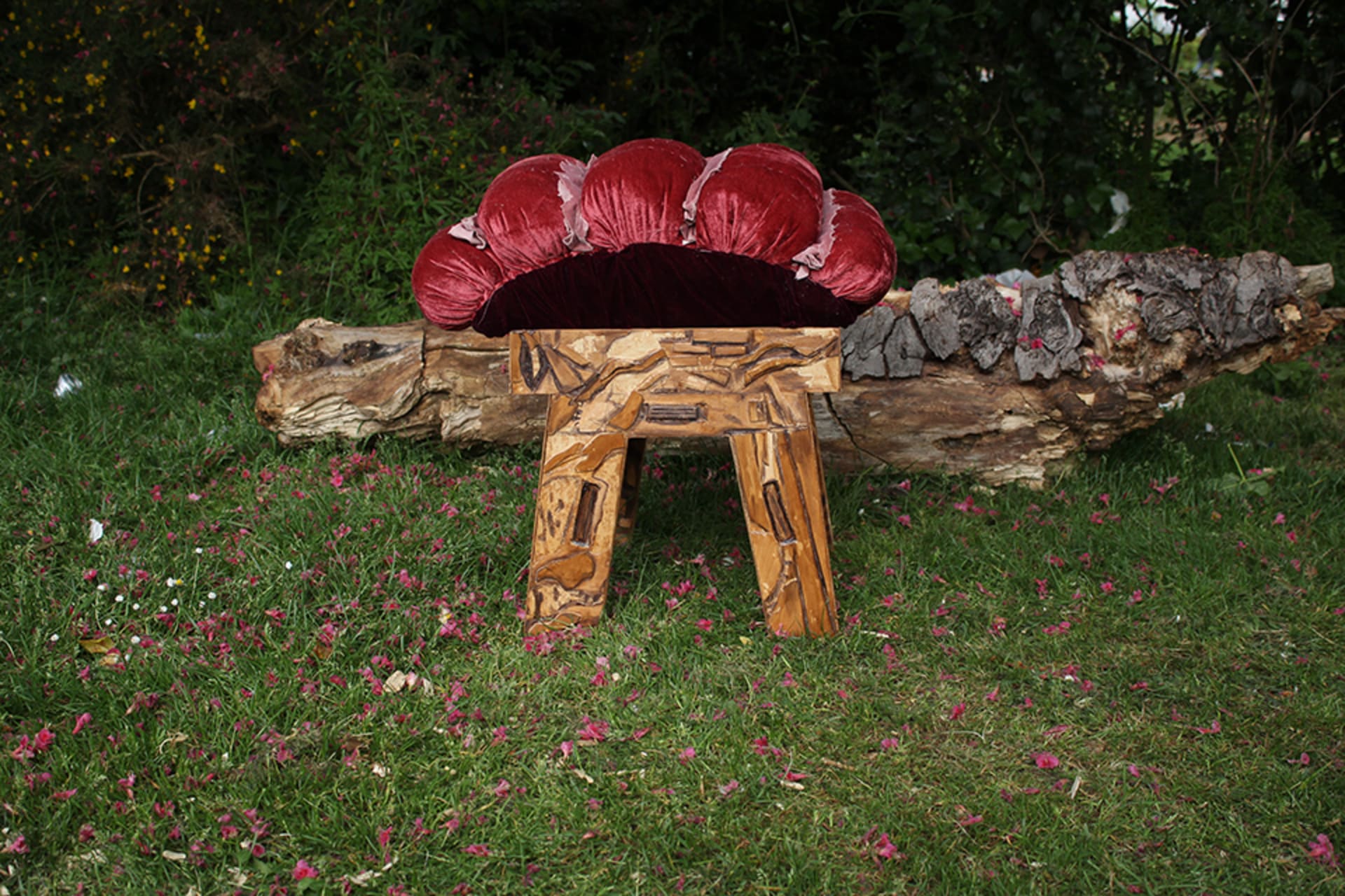 A small velvet upholstered stool, with a wooden hand carved base made to look like decaying wood. The stool is placed in nature infront of a dead log 