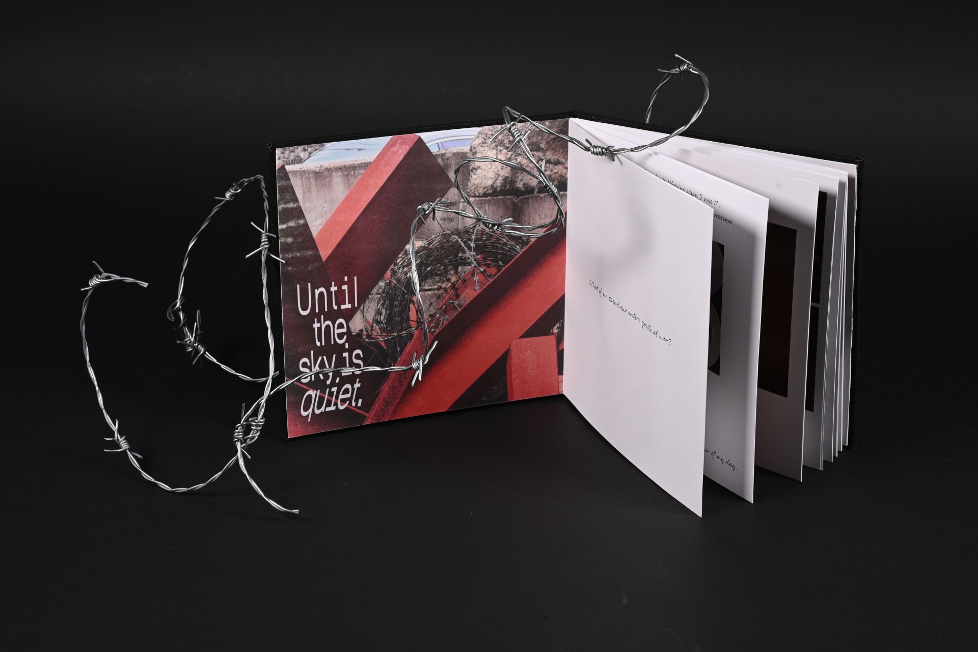 A handmade artist’s book lies open, wrapped in loose barbed wire. The cover features a photograph of red metal beams and reads “Until the sky is quiet