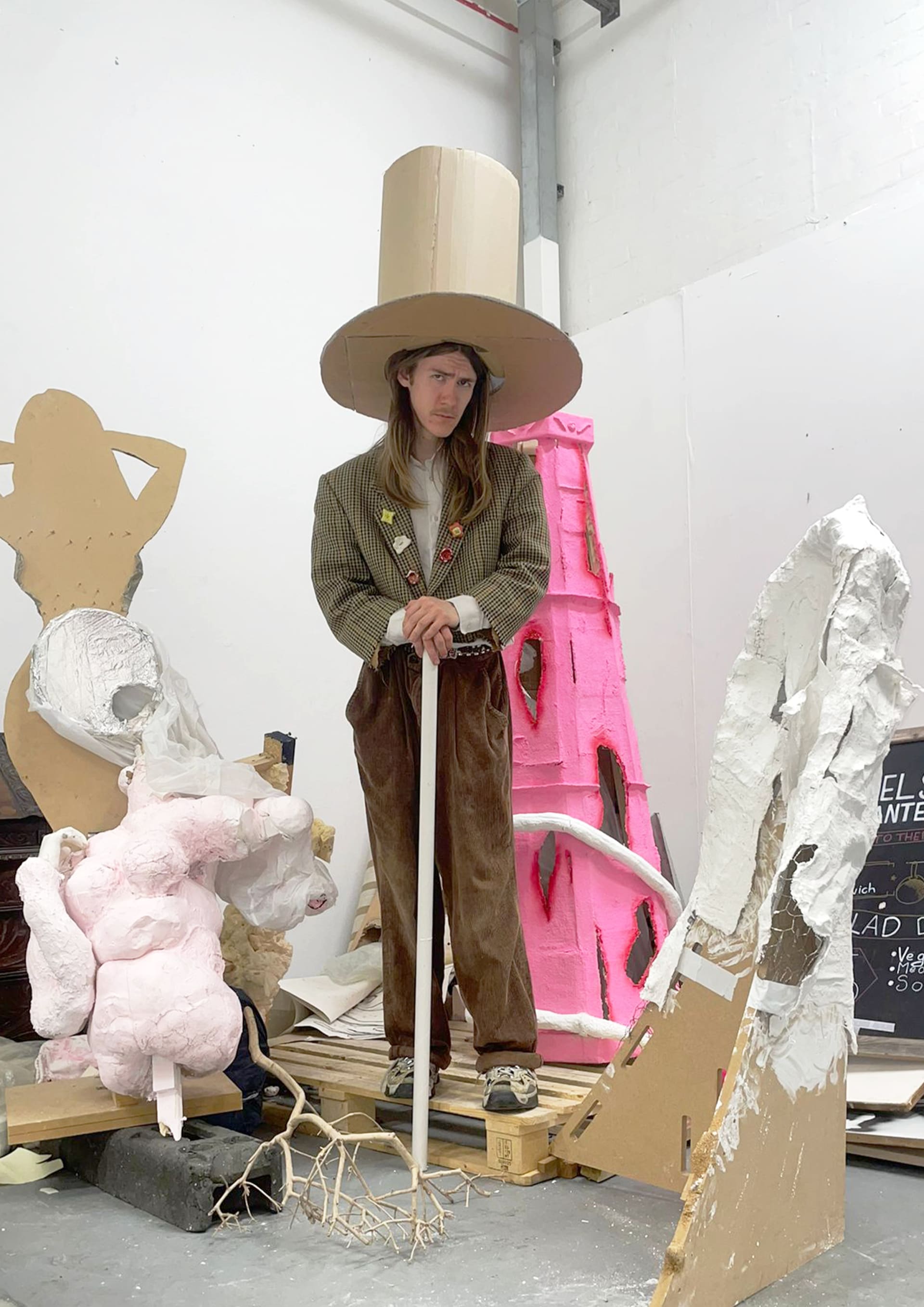 A man in a giant cardboard hat, stands amongst a pile of eclectic rubbish.