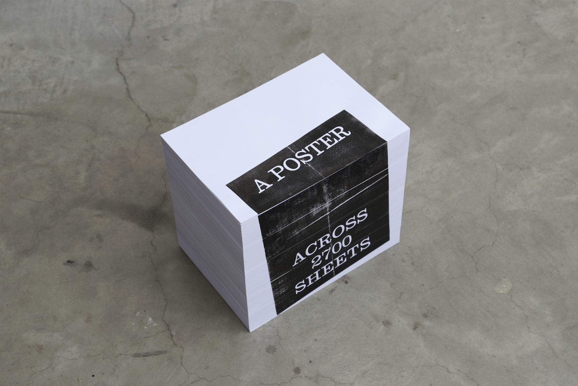 A thick stack of white paper sits on the floor. Along the side edges, black ink reveals part of a poster image, spread across 2,700 sheets.