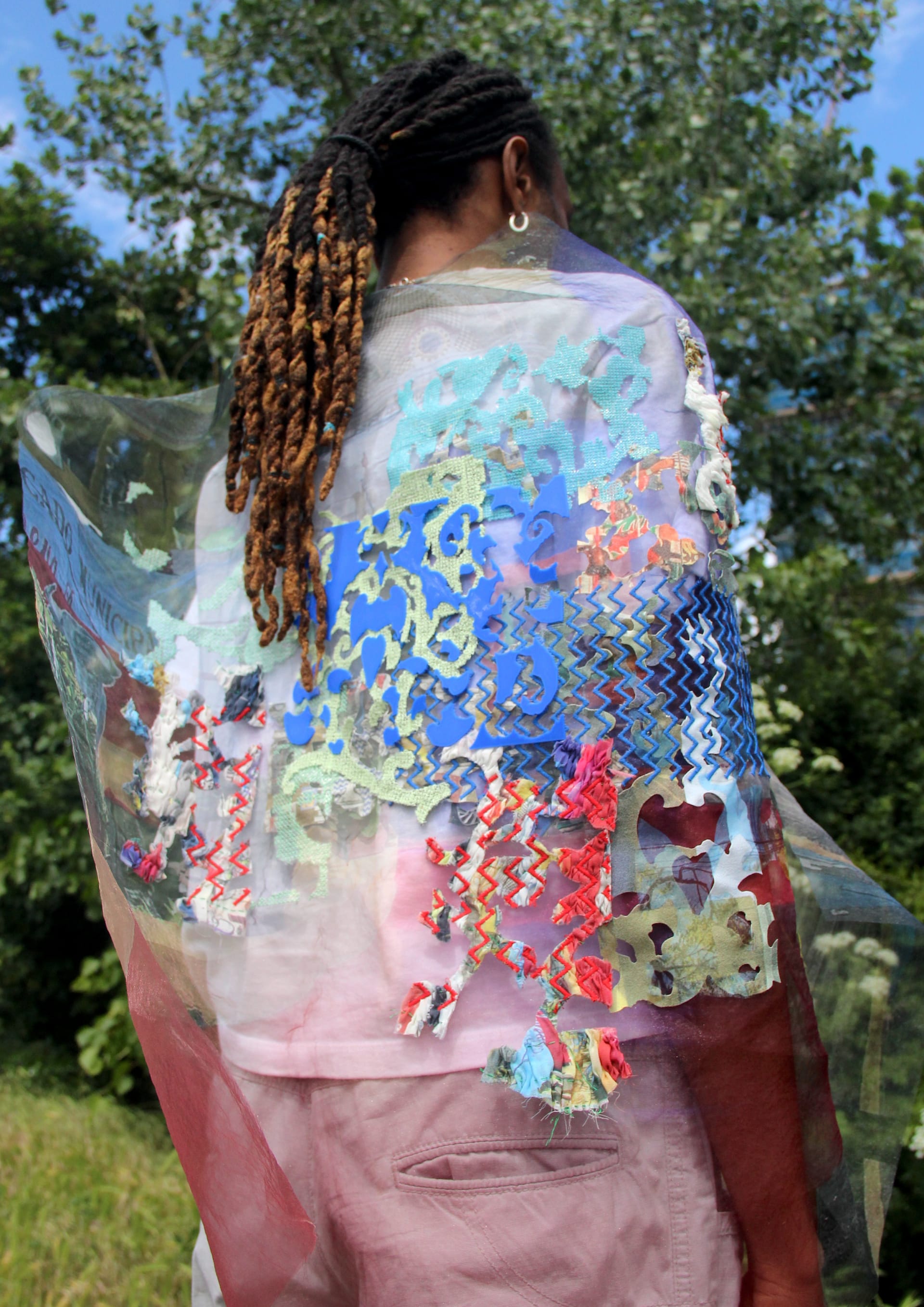 In the image the model, who is a young, black male is stood facing away from the camera. He is wearing all white with a colourful, but sheer fabric