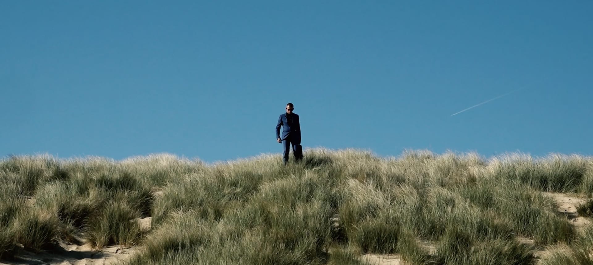 A man in a blue suit, stands on top of a grassy hill.