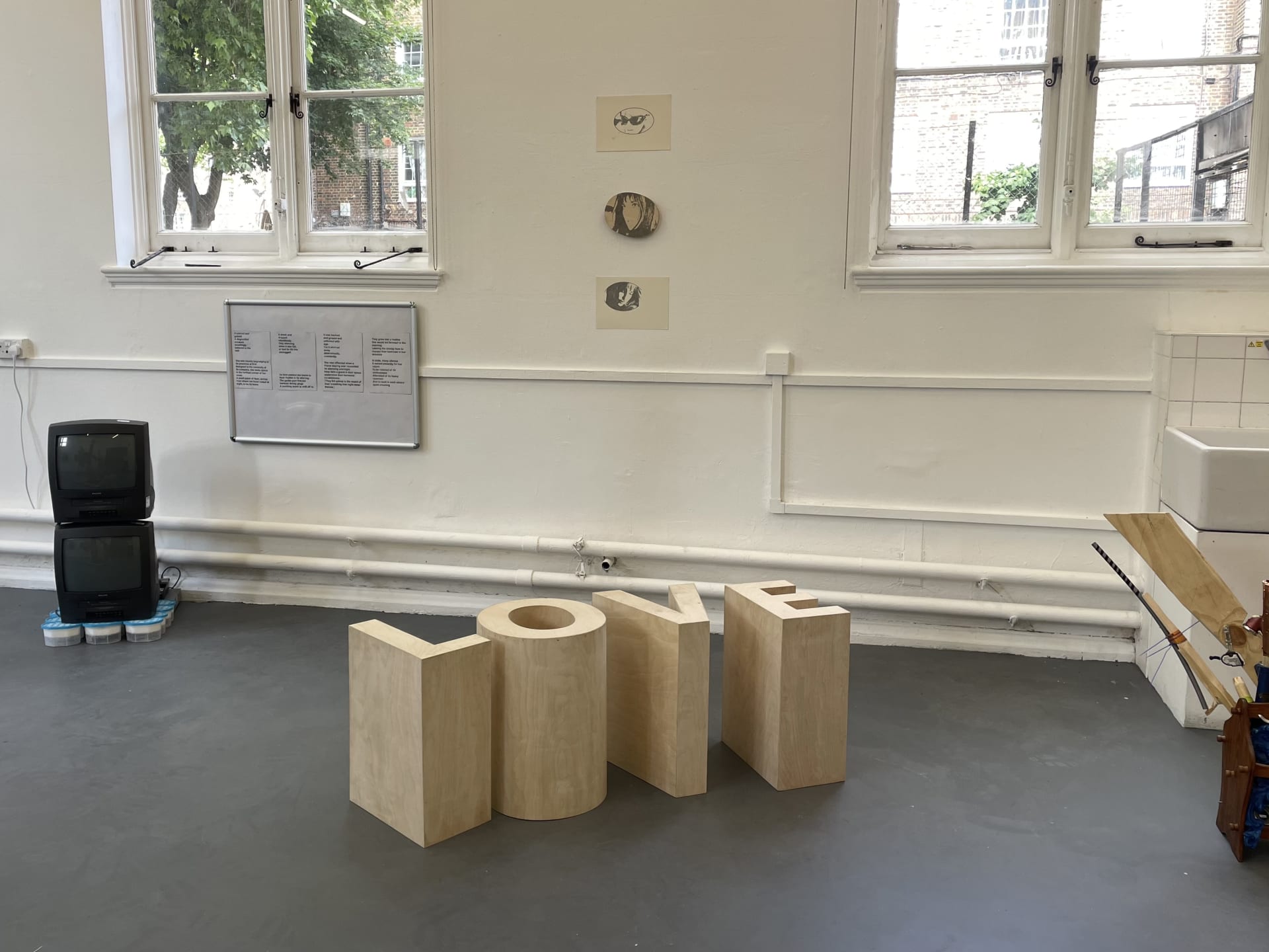 Landscape photo of gallery with wooden 3D version of the word 'love', horizontal stack of drawings, and surrounding works.
