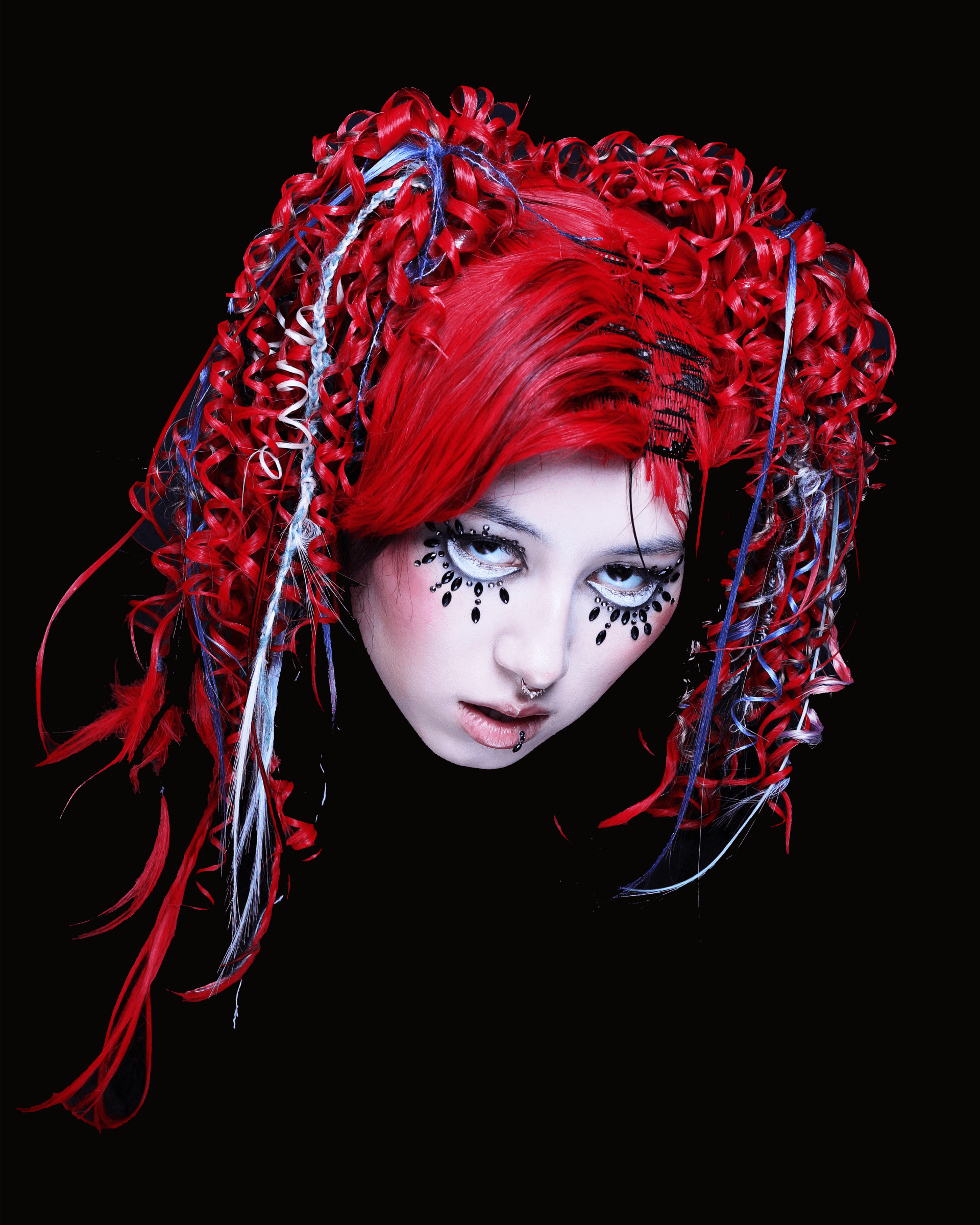 alt="A headshot of a person with bright crimson hair styled in elaborate, tightly coiled pigtails accented by blue and white strands, their pale face