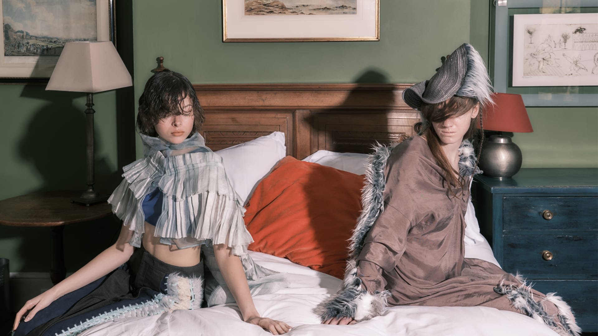 Two women are posed on a bed with a crisp white duvet and a bright orange pillow. Their expressions are serious.