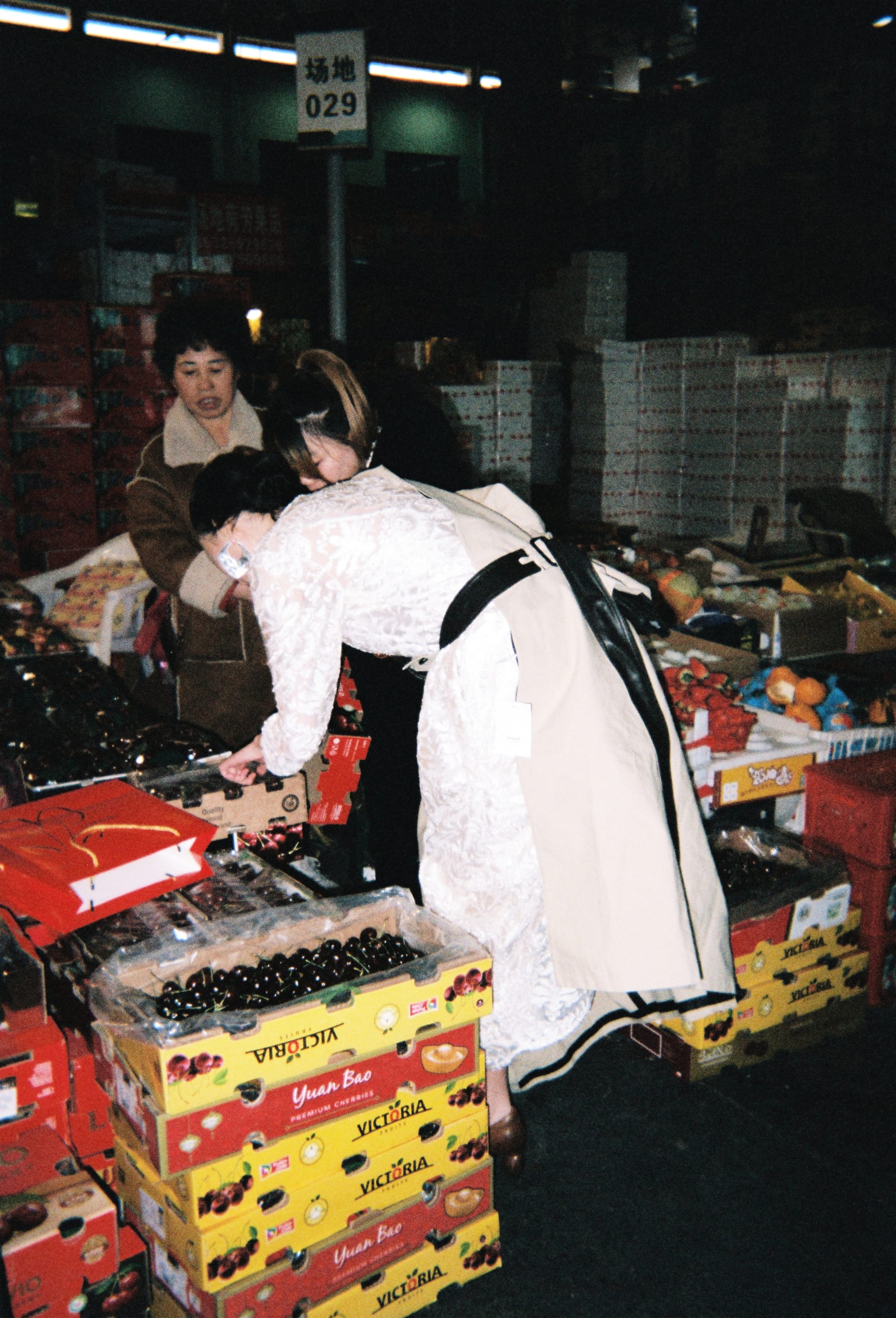 In a bustling local fruit market, a model in high-fashion clothing moves naturally among residents, selecting fruit. The scene creates a visual dialog