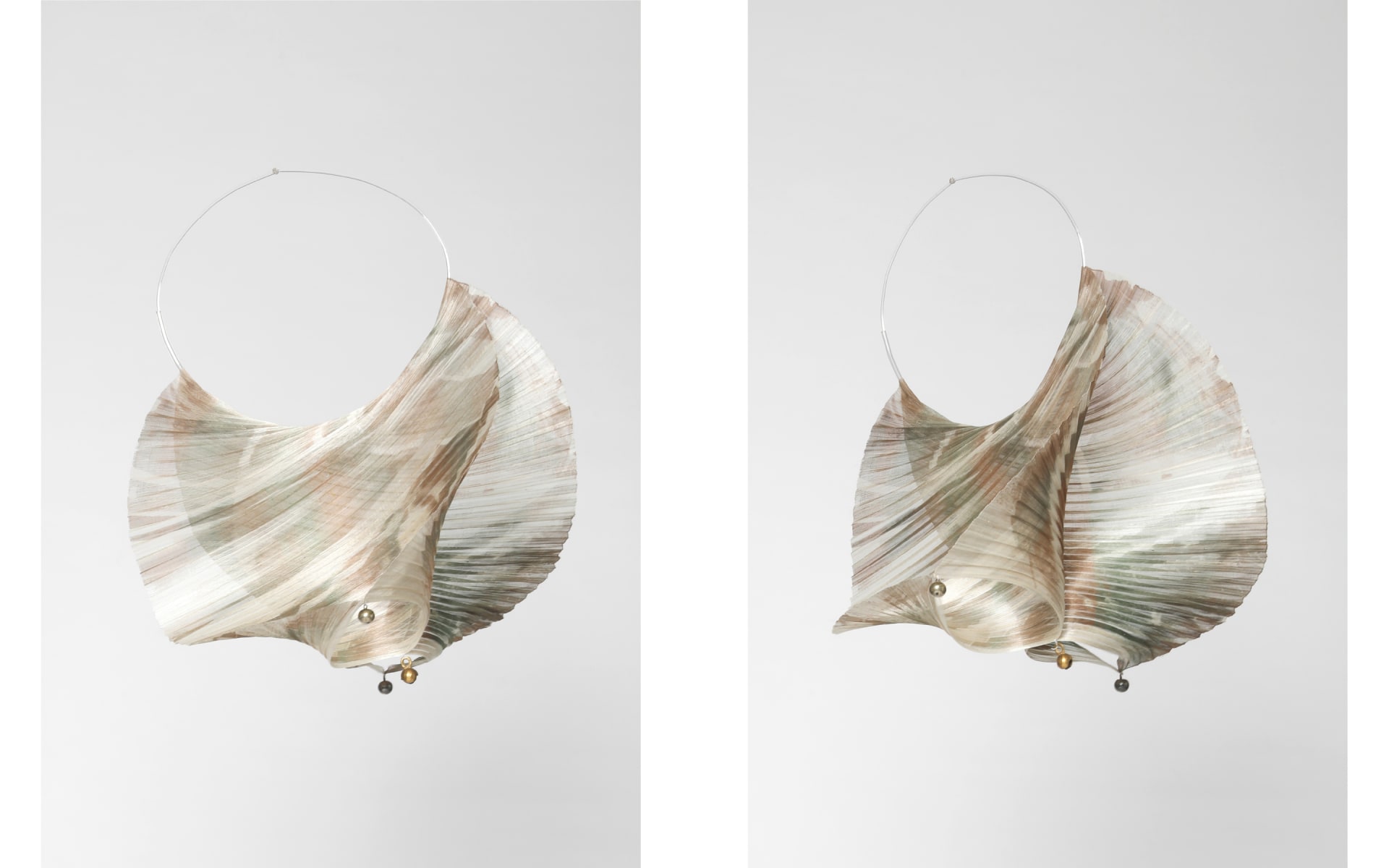 A lightweight, semi-transparent textile sculpture hangs from a thin circular wire. The textile is pleated and twisted into a fluid, shell-like form.