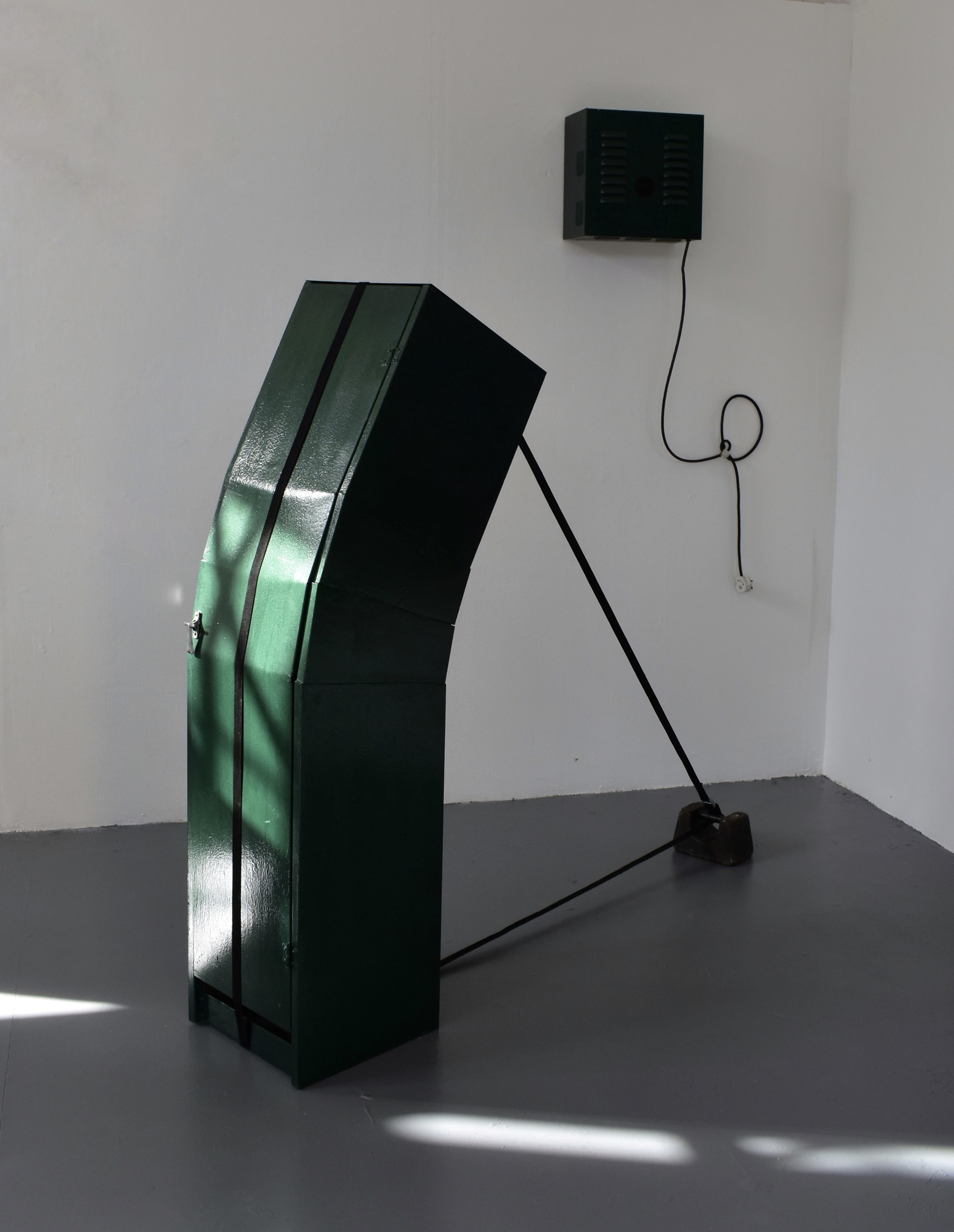 Green painted metal locker cut to bend into a curve, pulled by a black cam buckle strap attached to a cast iron weight on the floor.