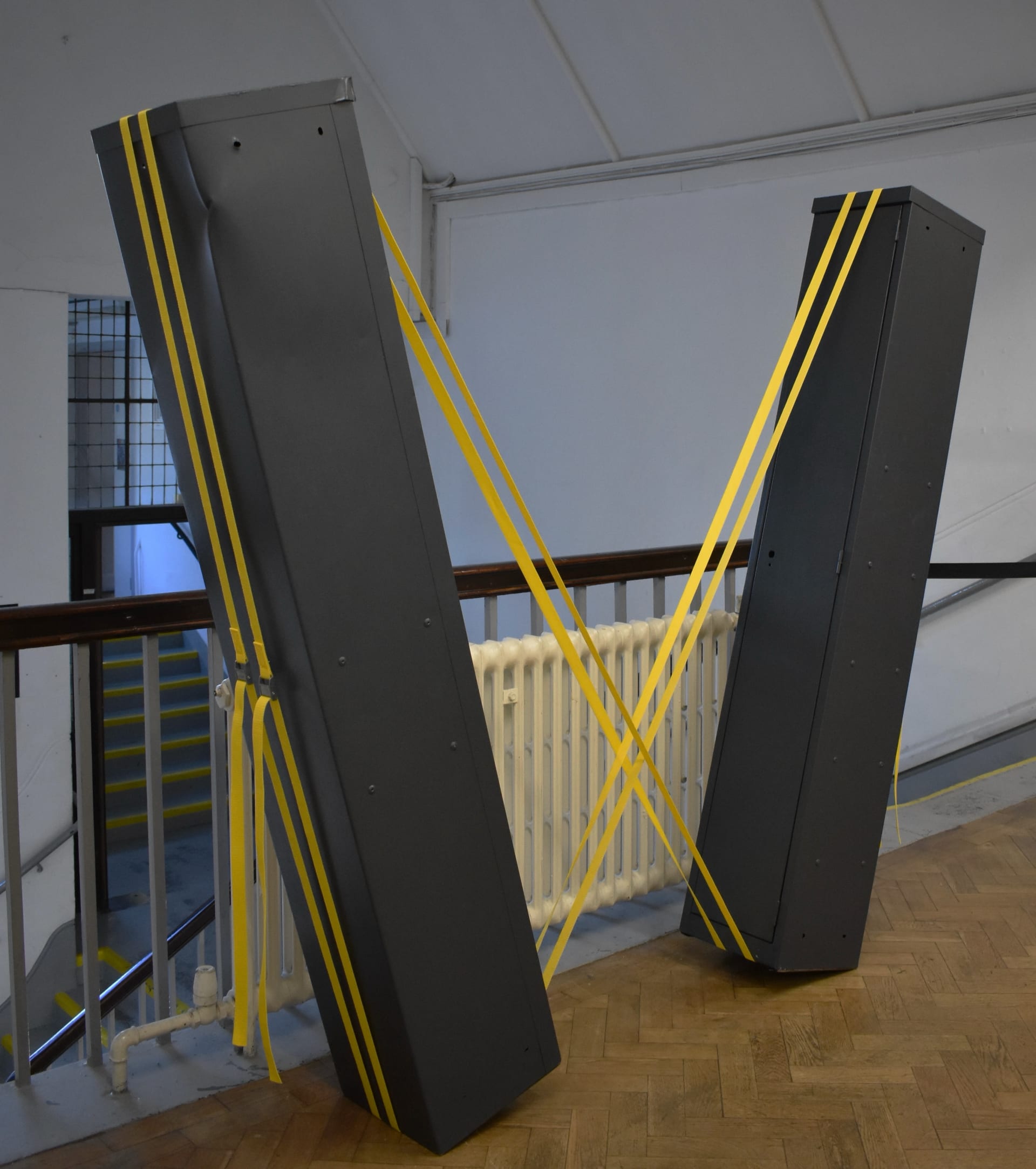 Two grey metal lockers, tilted and balanced, supported by two yellow cam buckle straps that cross over the lockers.
