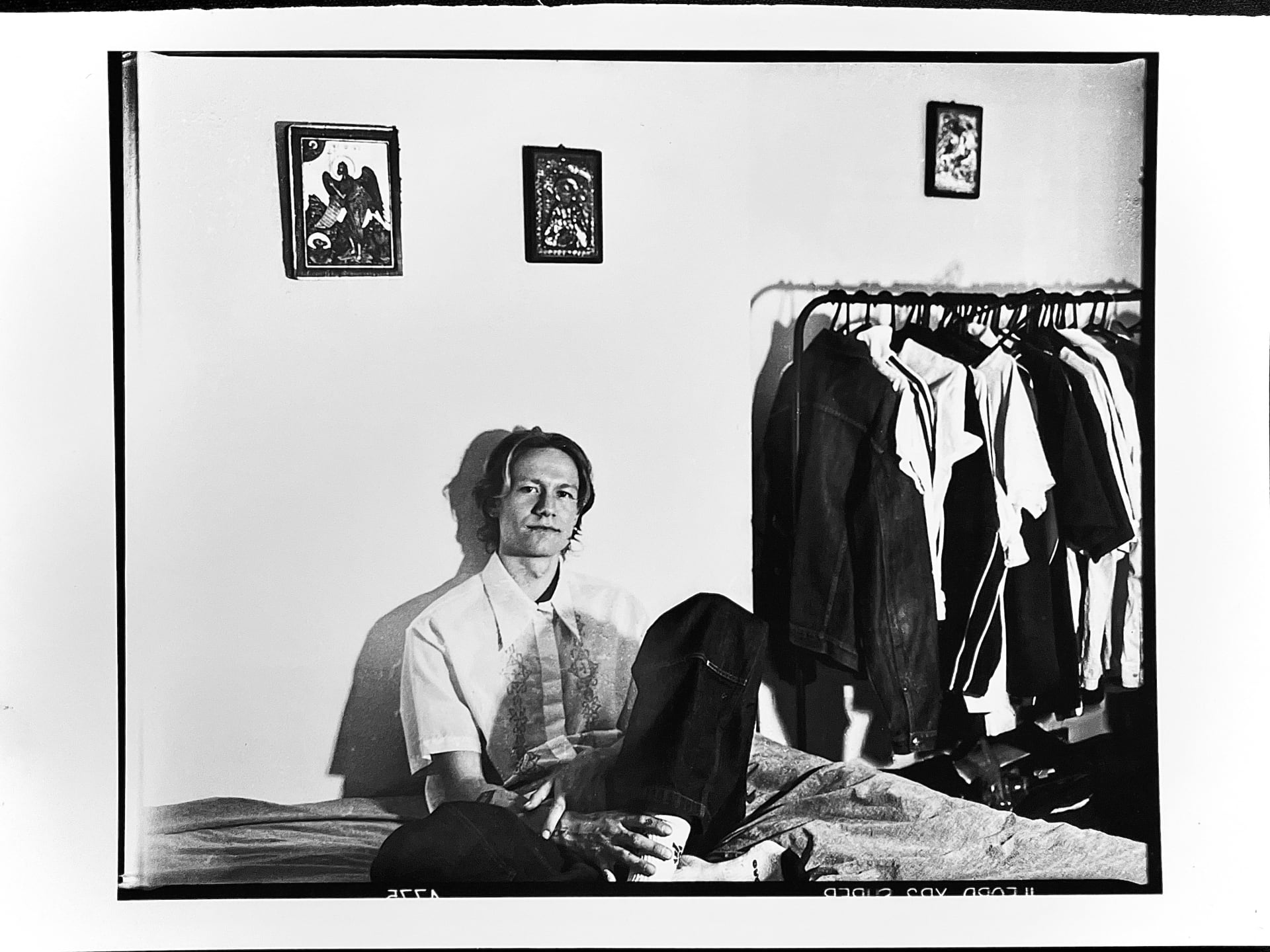 A black and white photo of a man sitting on a bed in a room. He wears a light shirt and looks calmly at the camera. Behind him, clothes hang neatly on