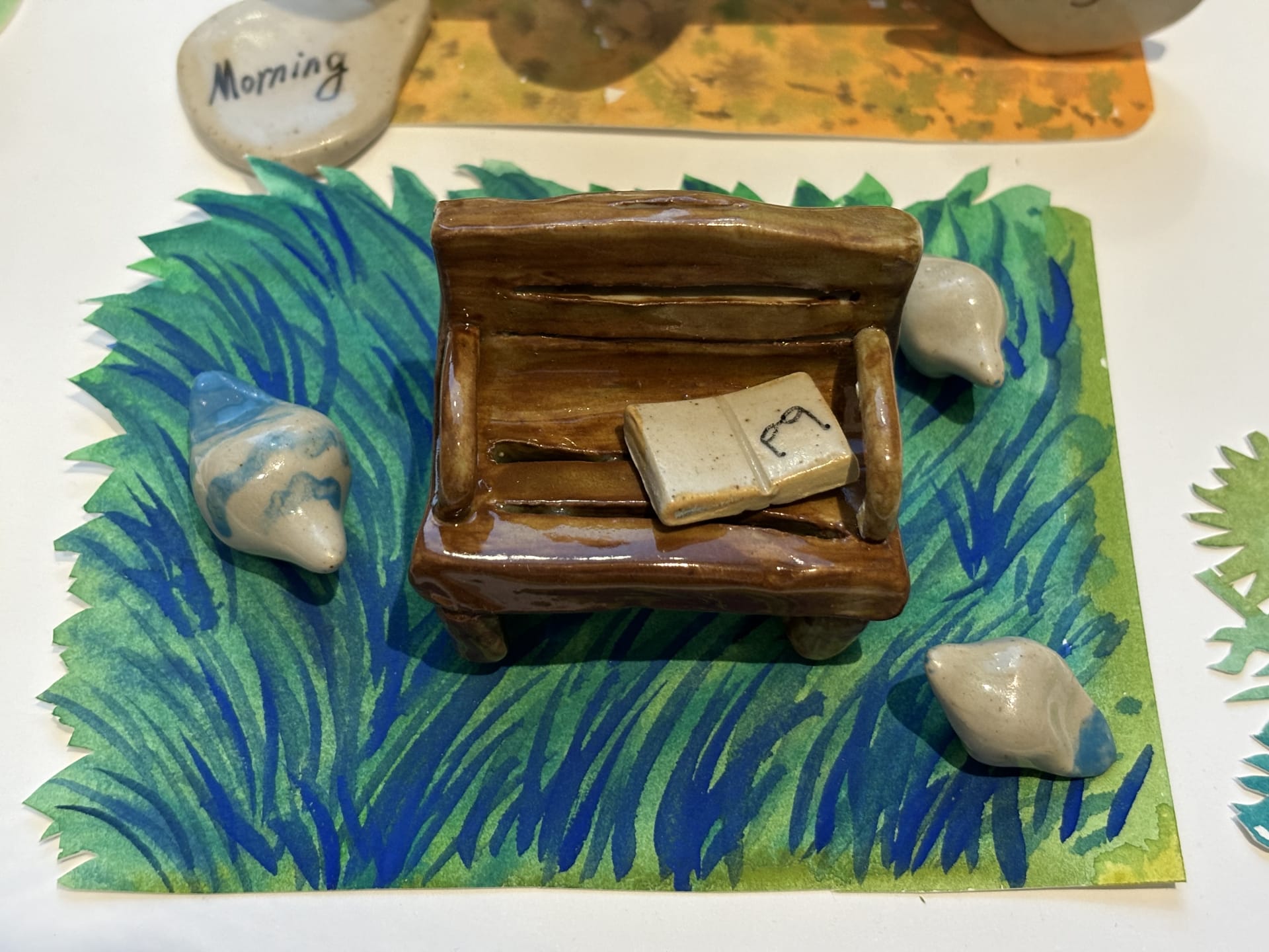 This image shows a small brown glazed ceramic bench placed on a piece of paper painted with green and blue grass patterns. On the bench lies an open c