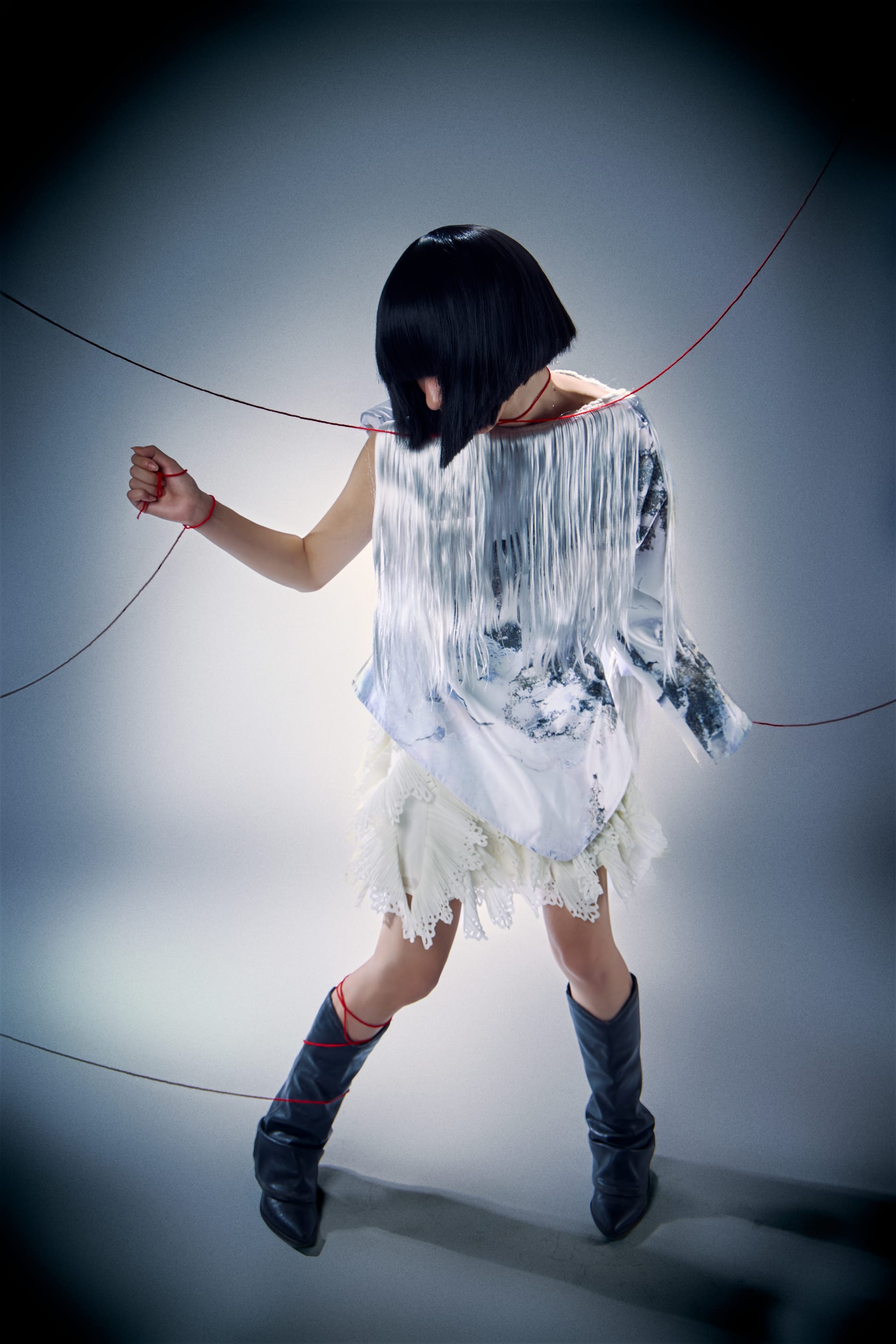 A figure with a short black bob is pulled by red strings, dressed in a fringed silver top and lace skirt.