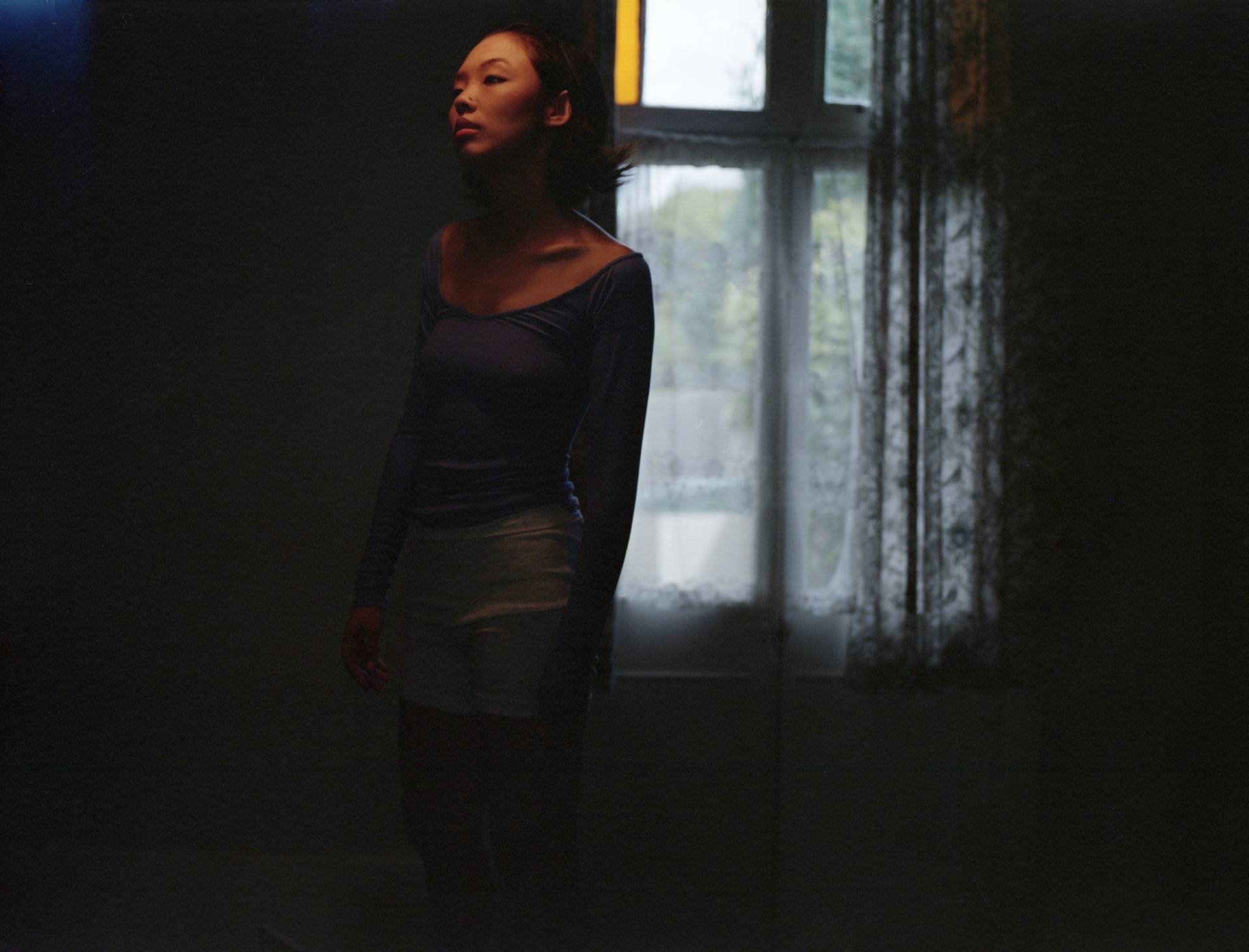 A person stands alone by a window in a dark room, wearing a blue top and white shorts, head slightly raised.