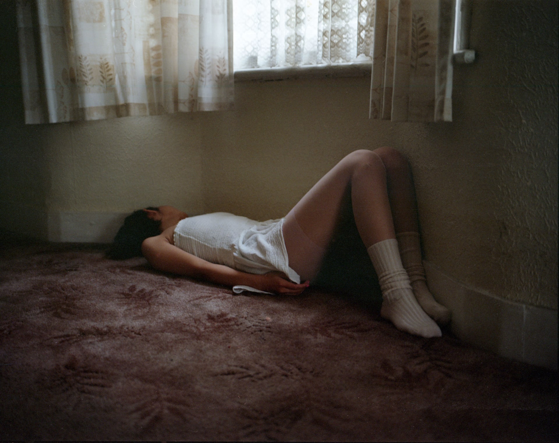 A person in a white dress and socks leans back in a corner on the carpet, faint light filtering through curtains.