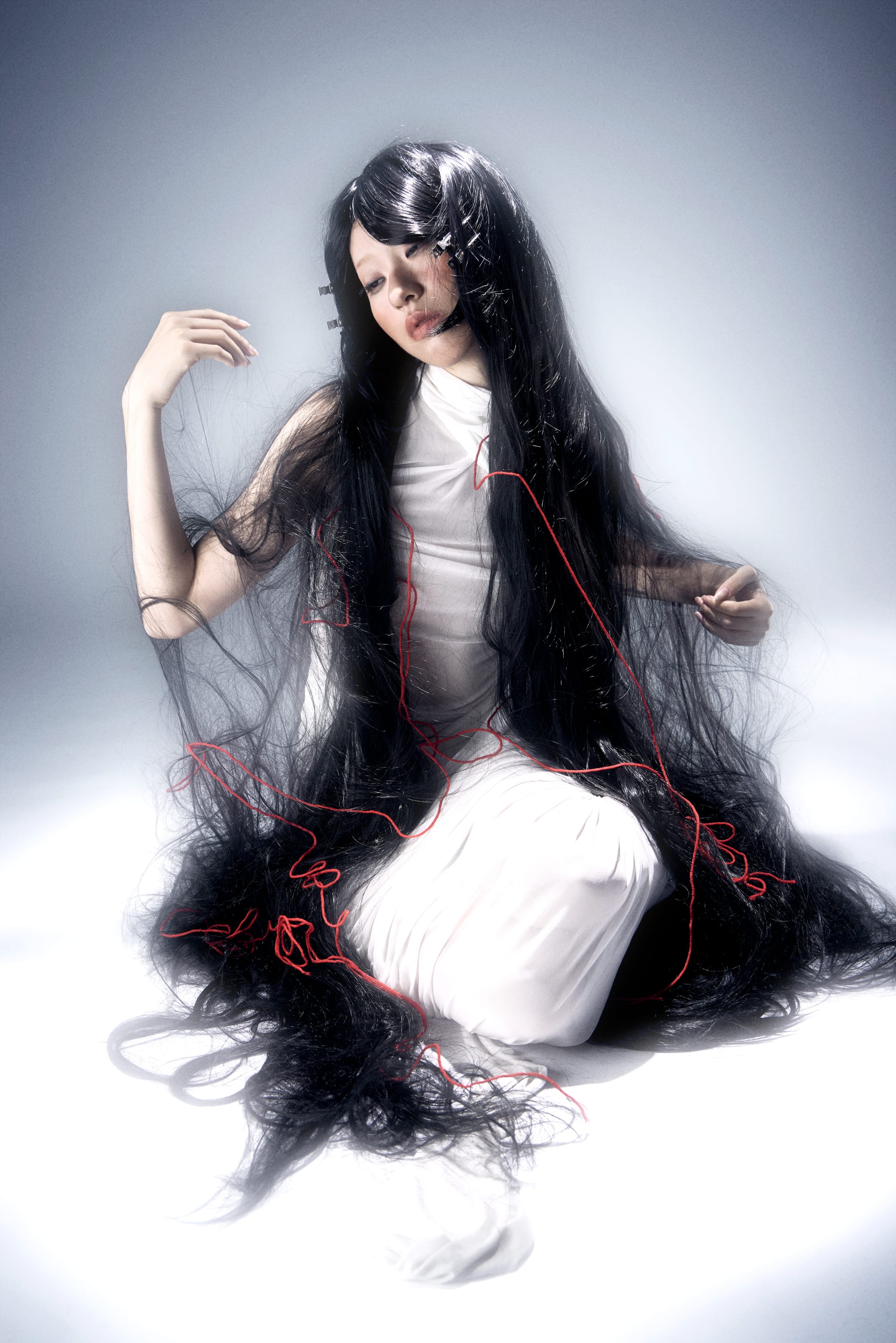 A woman in a long white dress sits with her eyes closed, draped in long black hair tangled with red threads.