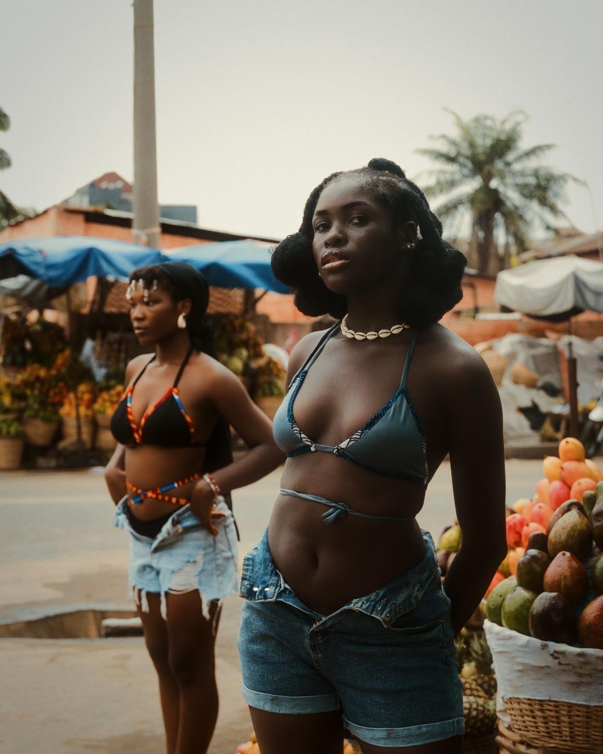 Two women modelling swimwear in denim shorts. They are standing in a market with fruit stalls behind them