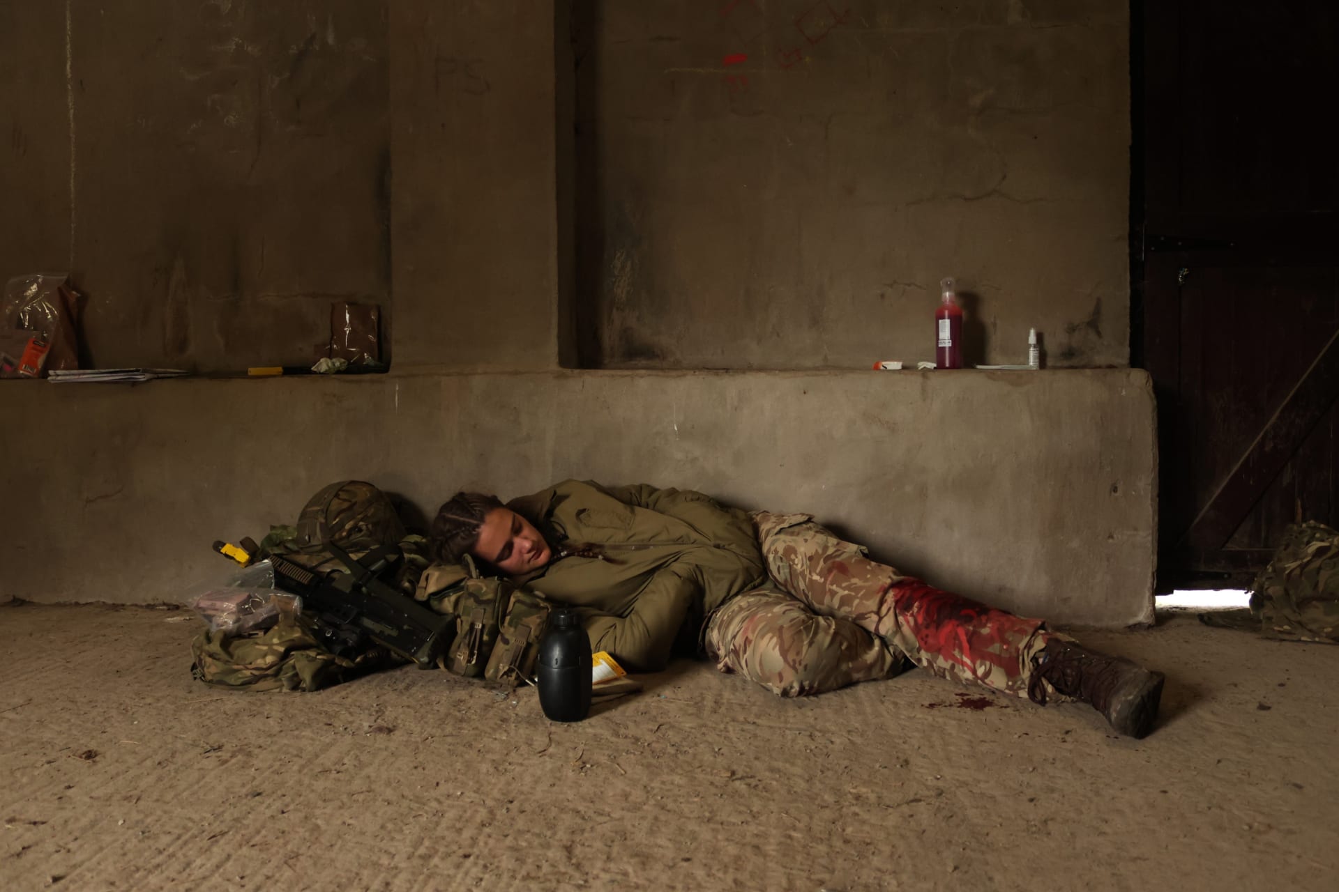 A young woman takes a nap. Resting her head on a small pile of her kit and with her rifle and water bottle resting next to her.