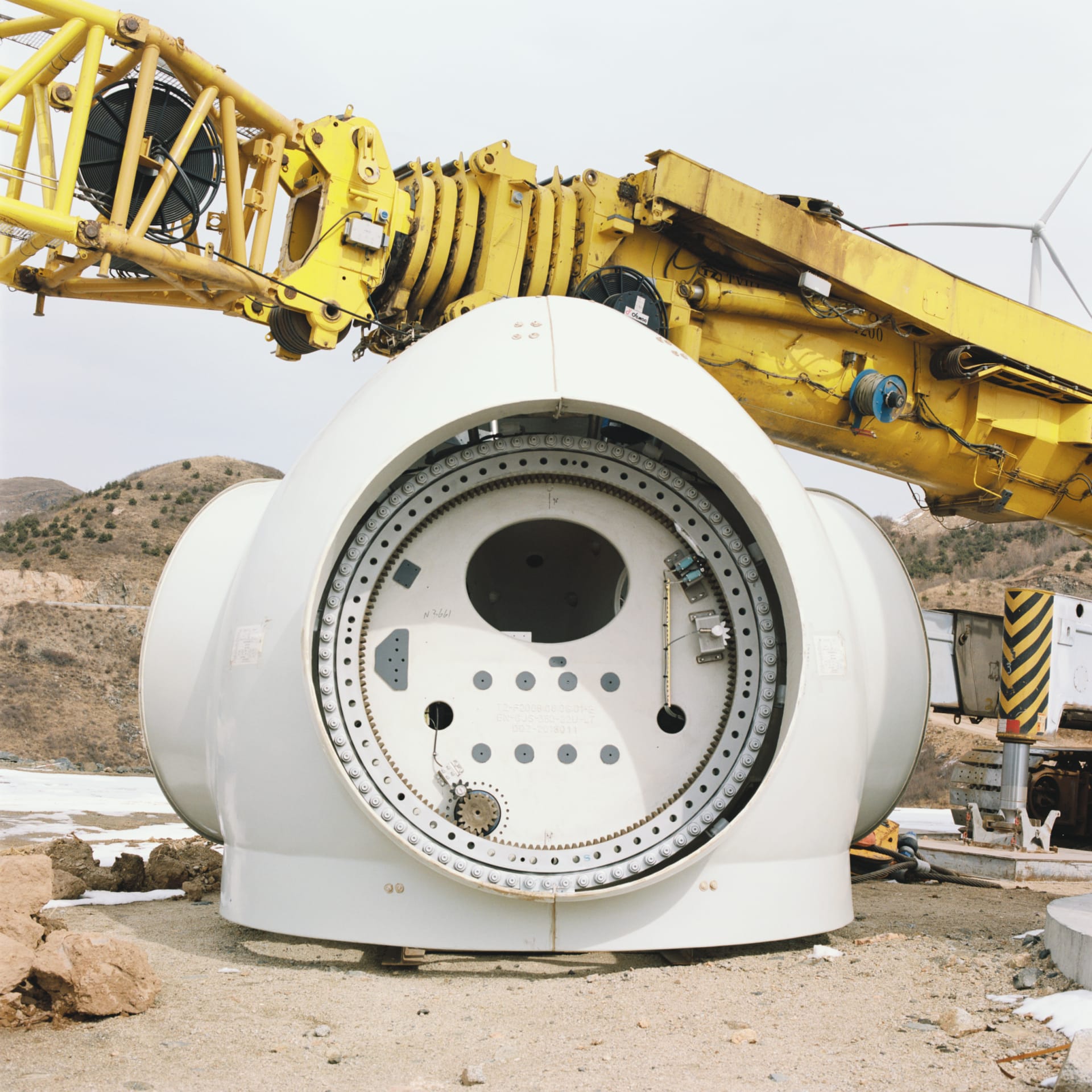 A large white wind turbine nacelle beside a yellow crane on a construction site.