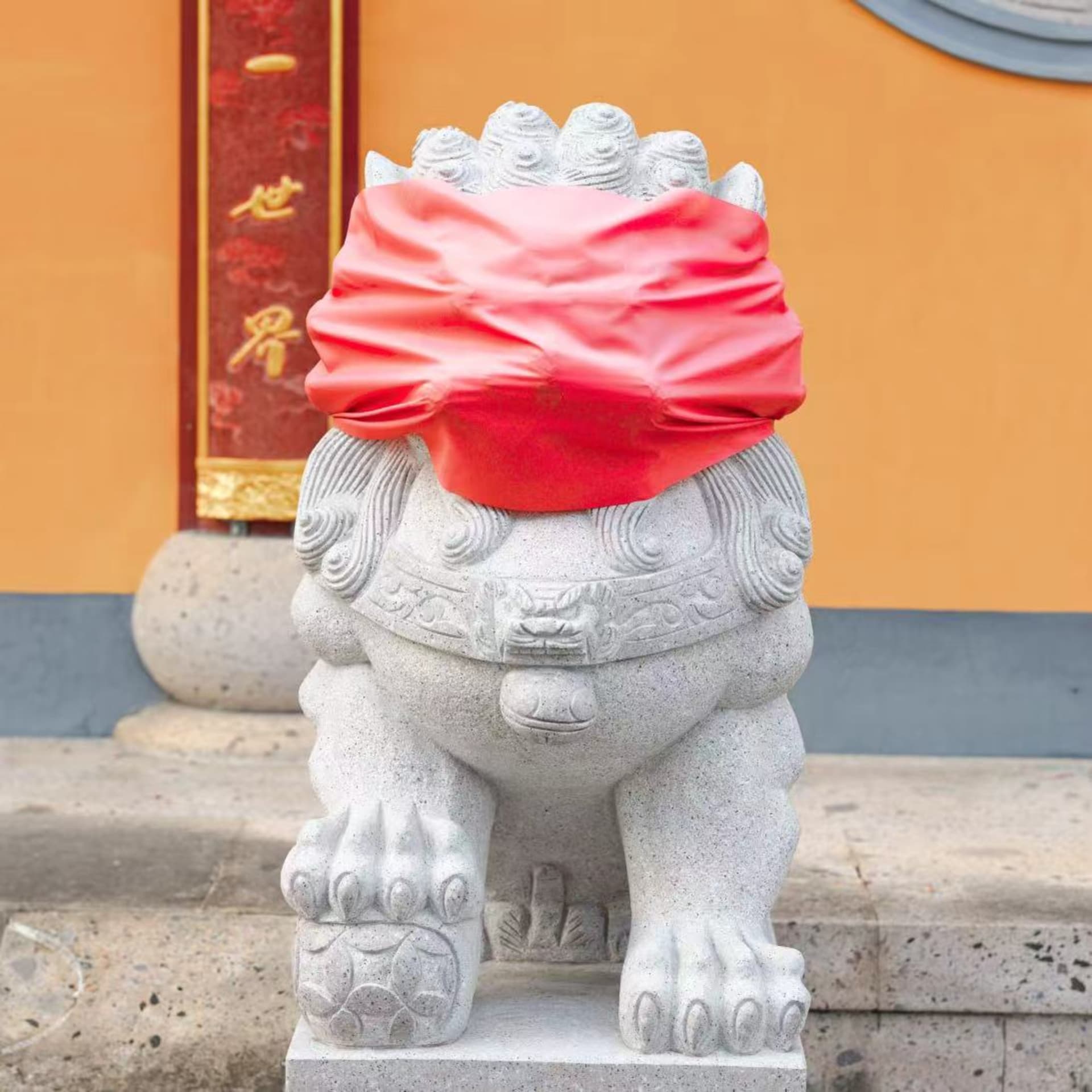 A stone lion statue had its eyes covered with red cloth, with the temple it stood in visible behind it.