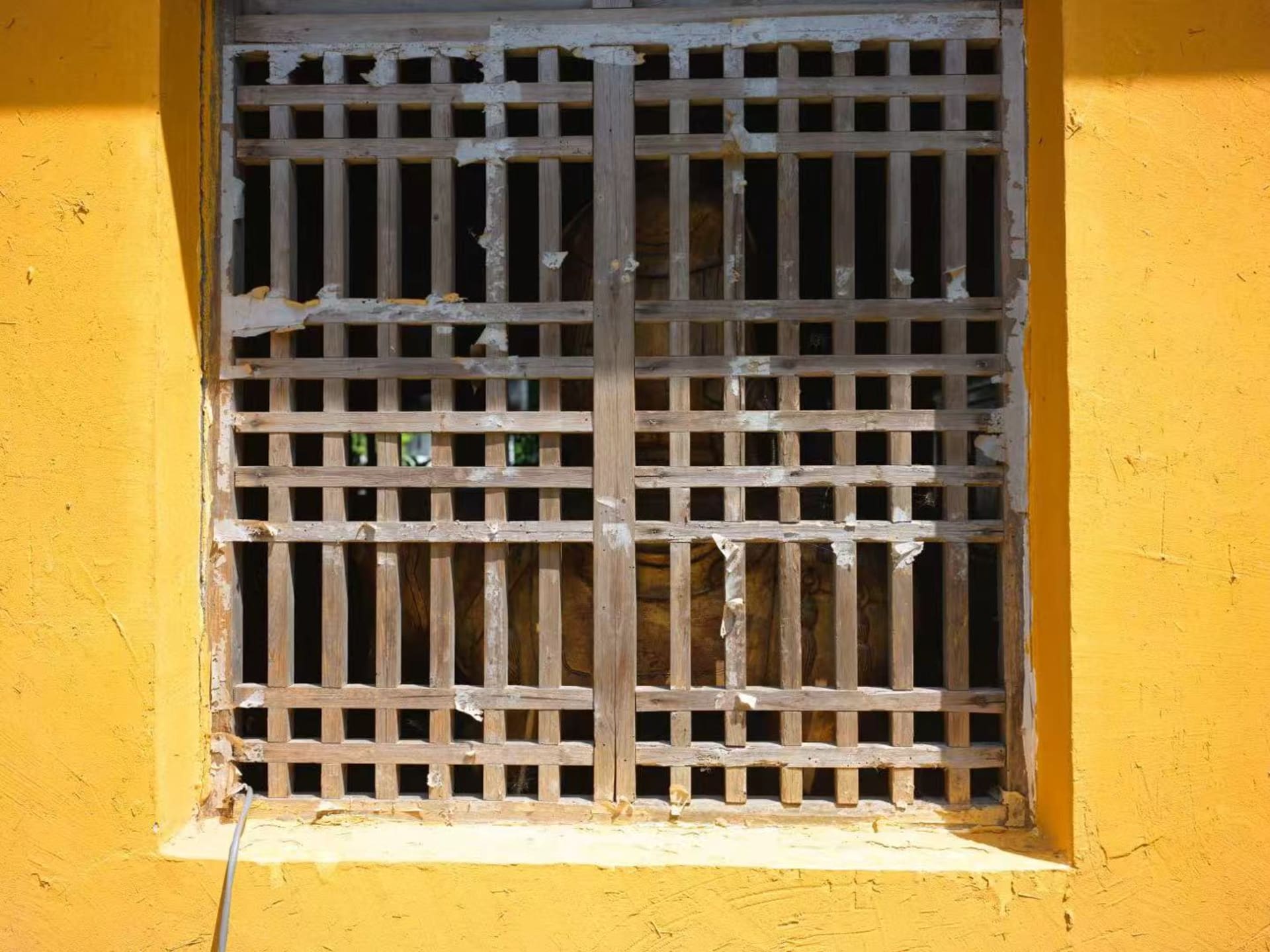 A yellow wall featured a window framed with wooden slats. Through this window, one could see a room beyond the wall, where a massive Buddha statue.