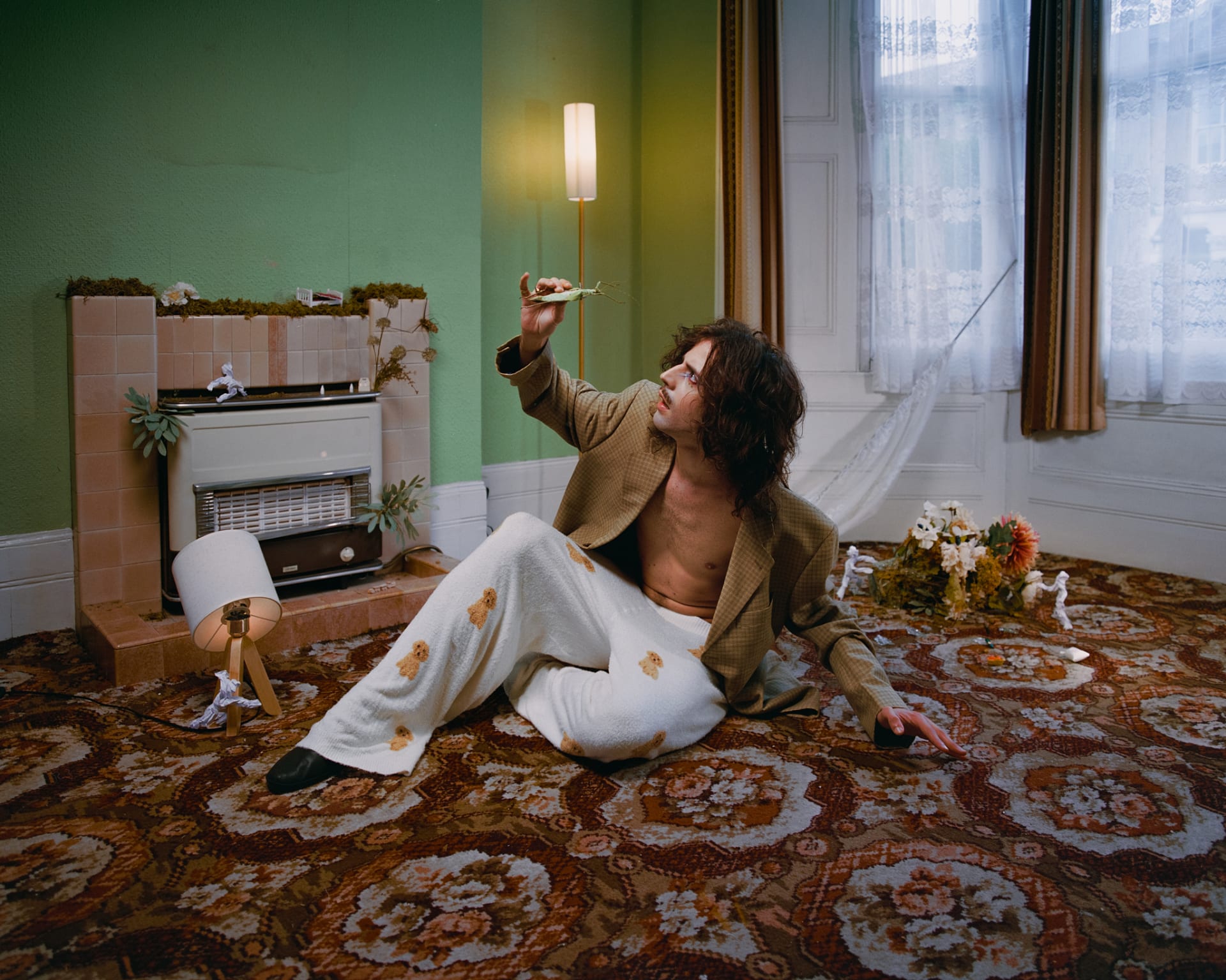 A man sits on a patterned carpet in a retro living room, gently holding insect in his hand.