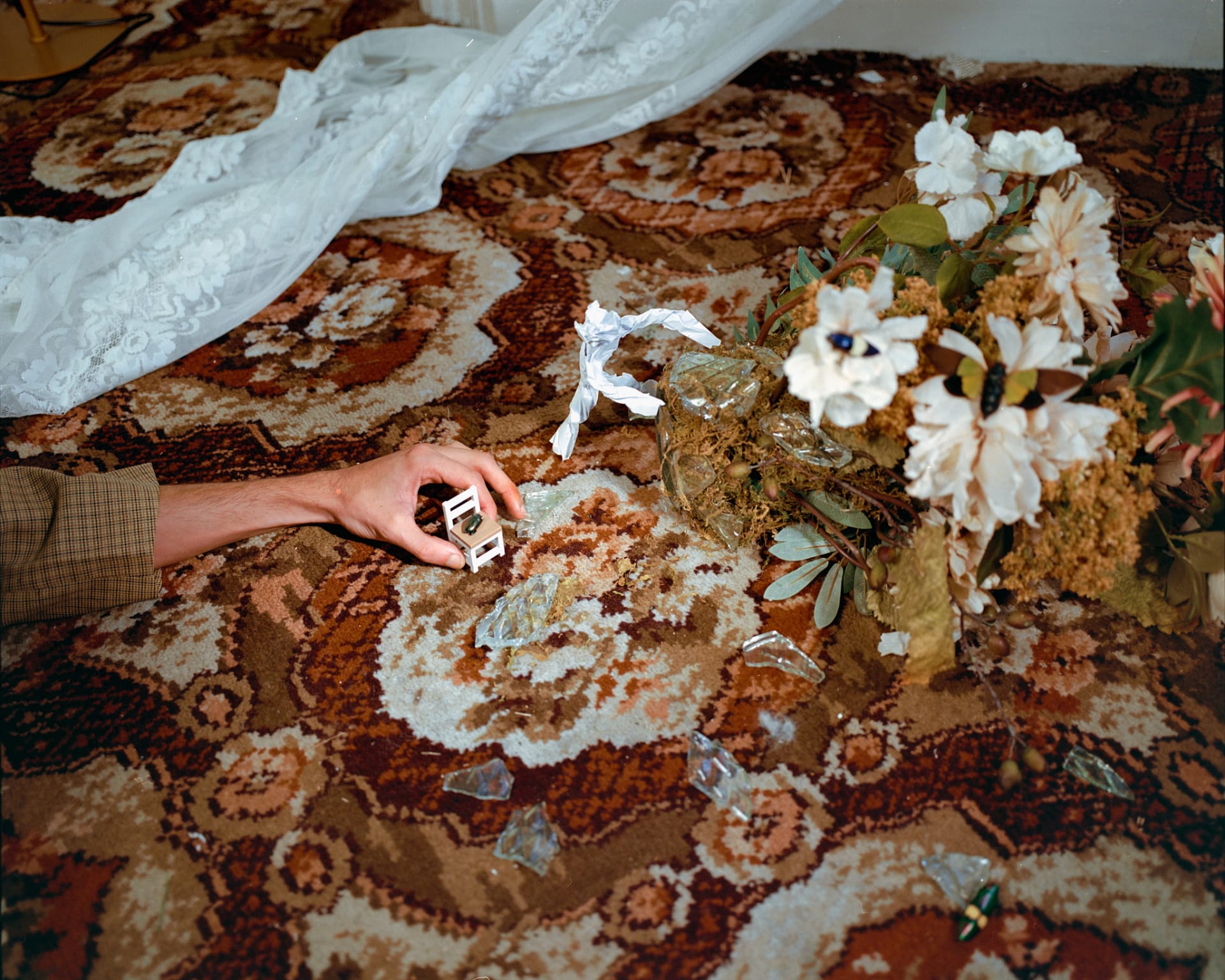 A hand reaches across a patterned carpet toward a small paper chair, surrounded by broken glass, flowers, and insects.