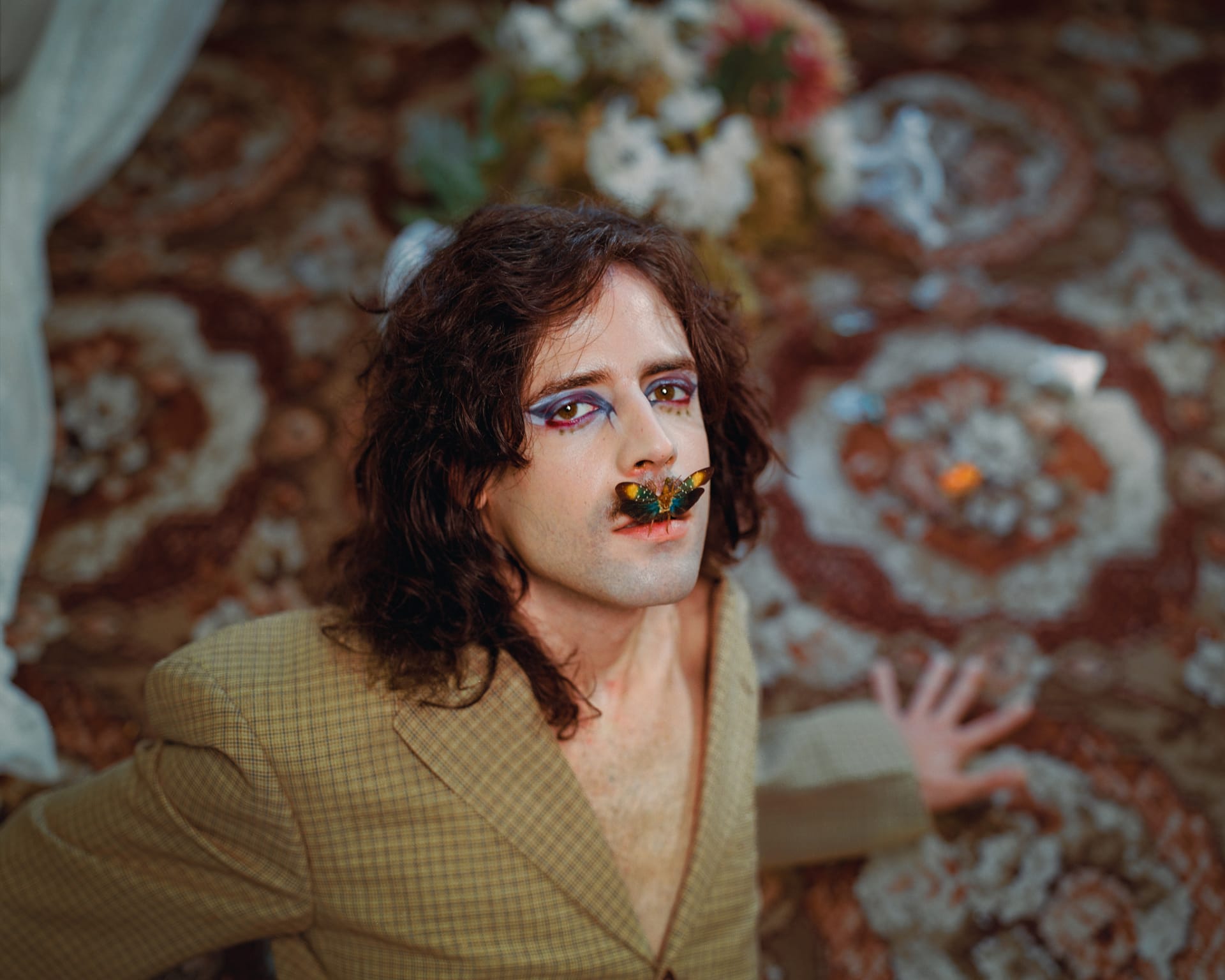 A man with long hair and makeup sits on a patterned carpet with a large beetle resting on his lips.