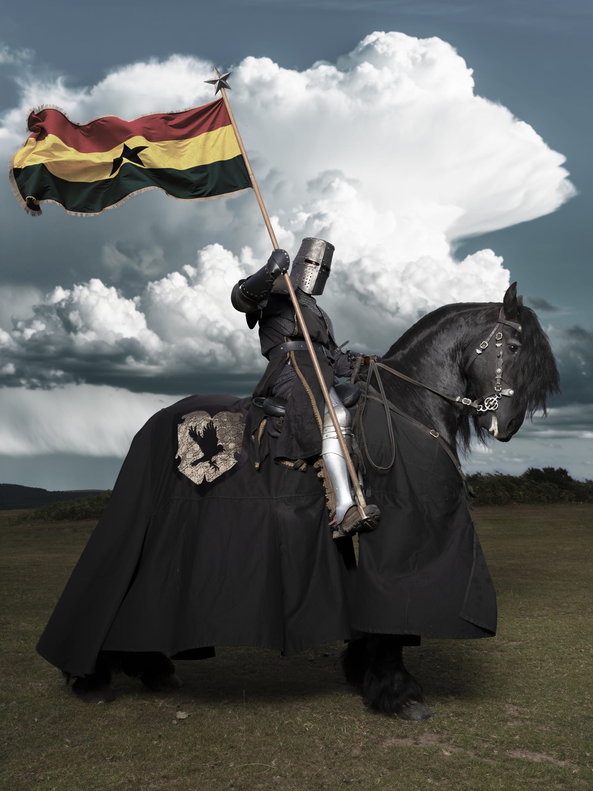 A Knight sits upon a black horse, in a green field. Grand clouds fill the sky and in his hand is a ghanaian flag on a pole supported by his boot.