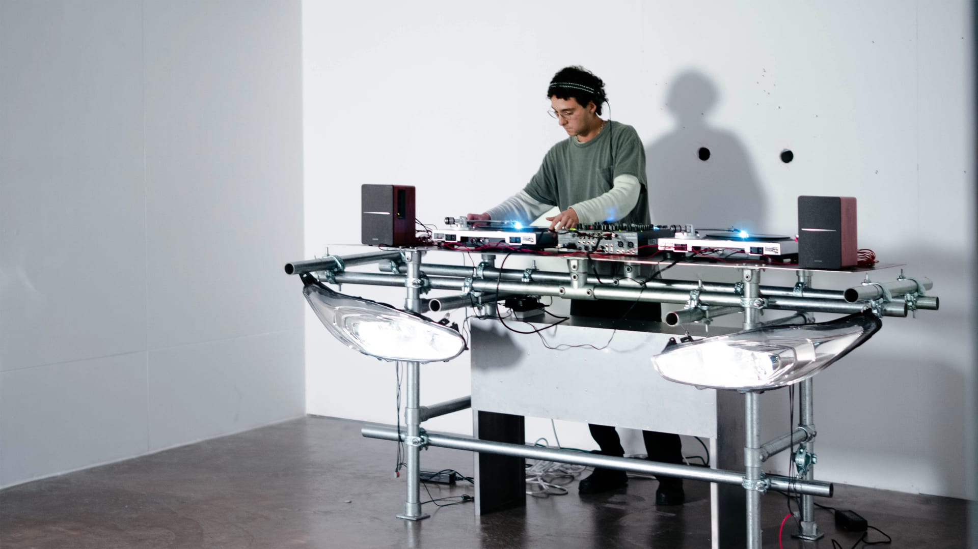 A metal pipe DJ table stands with car headlights hanging at the front. A DJ is playing on the equipment set on top.