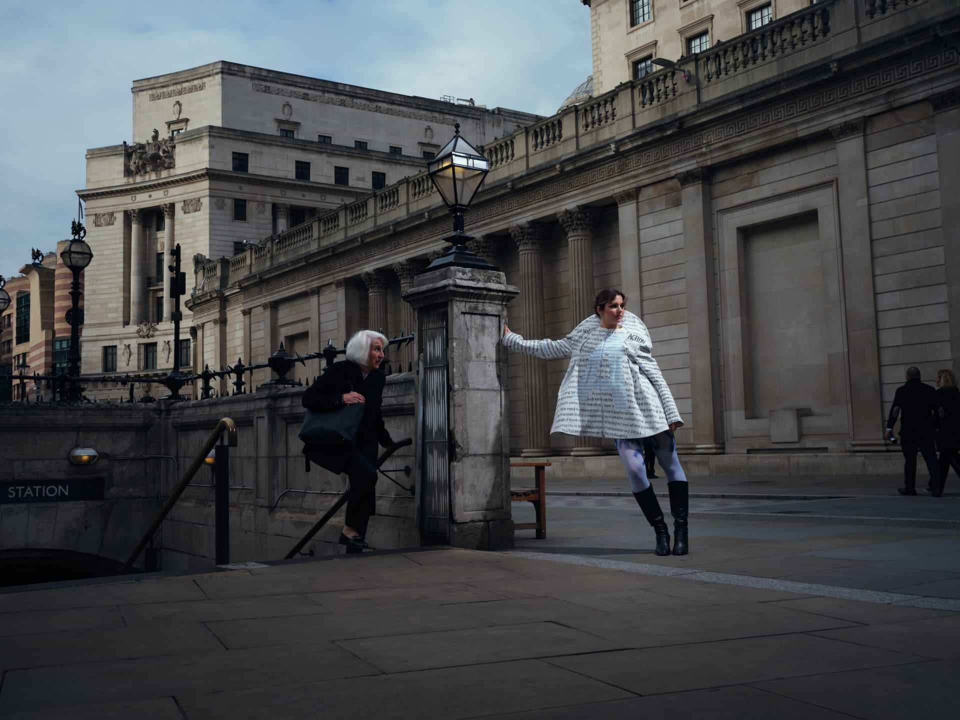 Senior lady walking out of a tube station and looking at the model wearing the hand-printed Accountable Jacket, shot in governmental district