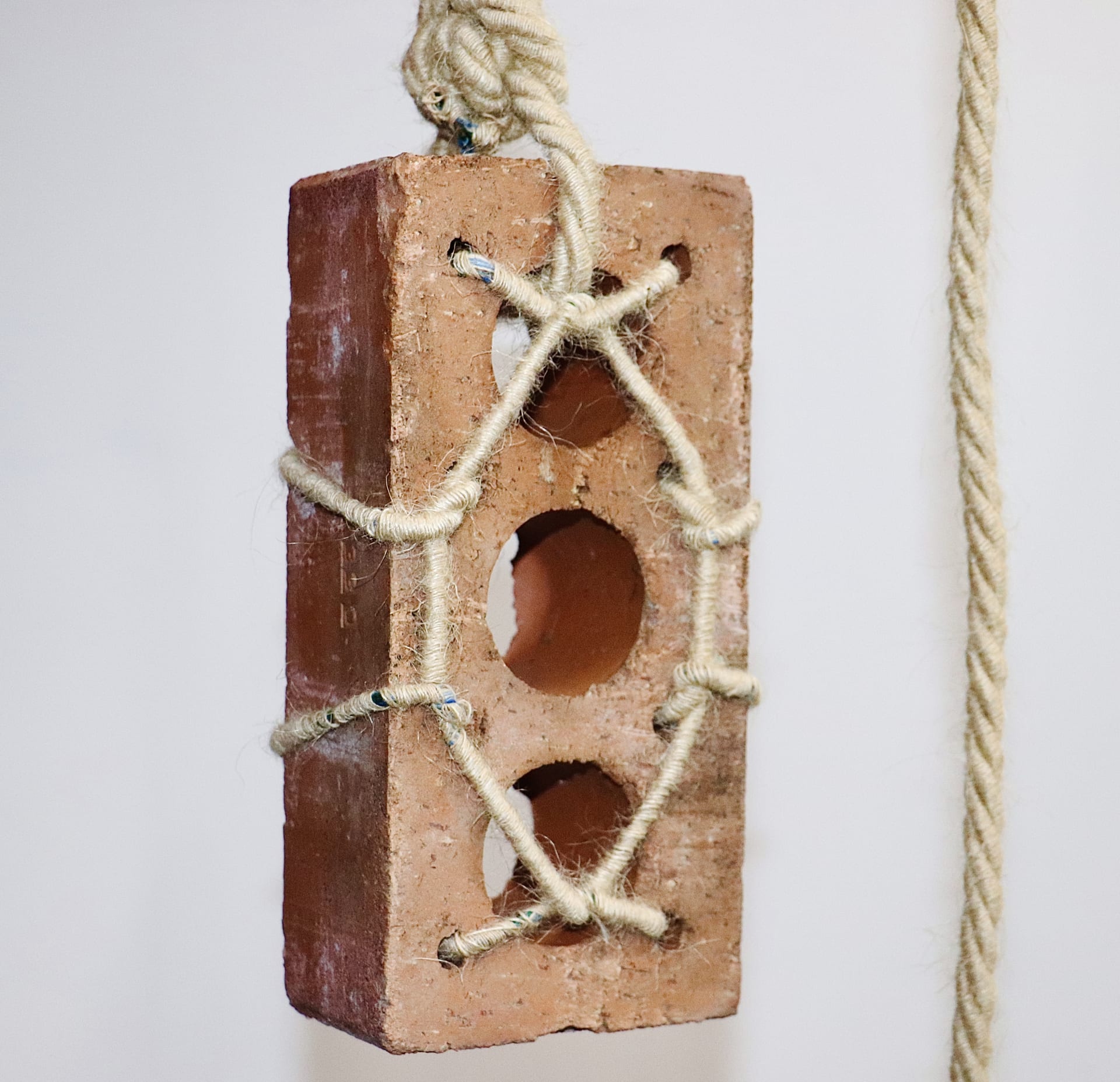 Red brick suspended in rope. Three large holes vertically in brick and four small holes on the edges, rope threaded through in bondage-like pattern.