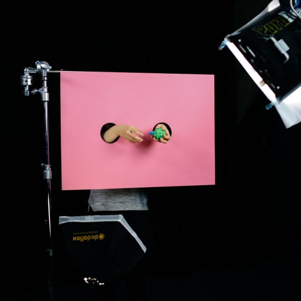 Two hands emerging through hole in a pink screen holding a green item