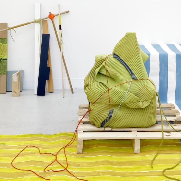 A room shot of colourful fabrics laid across the floor. There is a bean bag structure wrapped in red string sitting on a pallet