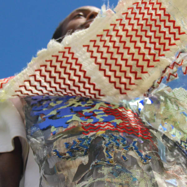 Person wearing a vibrant, abstract outfit with metallic textures and zig-zag patterns, set against a clear blue sky. The mood is bold and artistic.