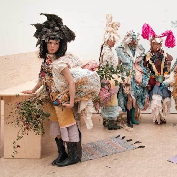 Models in avantgarde garments in muted and pastel colours are posing backstage. All are wearing headdresses and there are woven and plaited textiles and elements, patterns and tree branches incorporated.